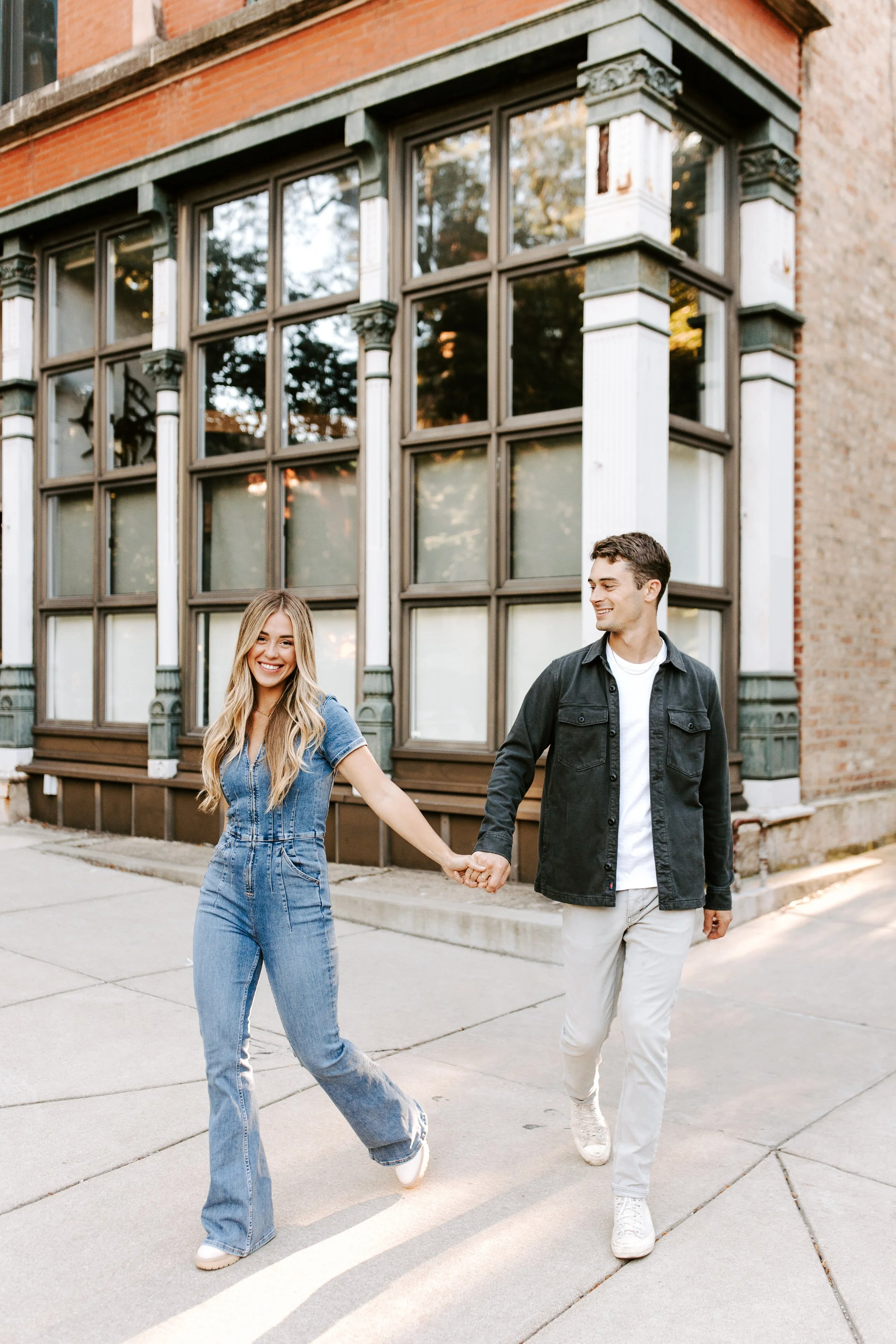 A young woman and a young man holding hands while walking on a city sidewalk in front of a large brick building with decorative architectural details.