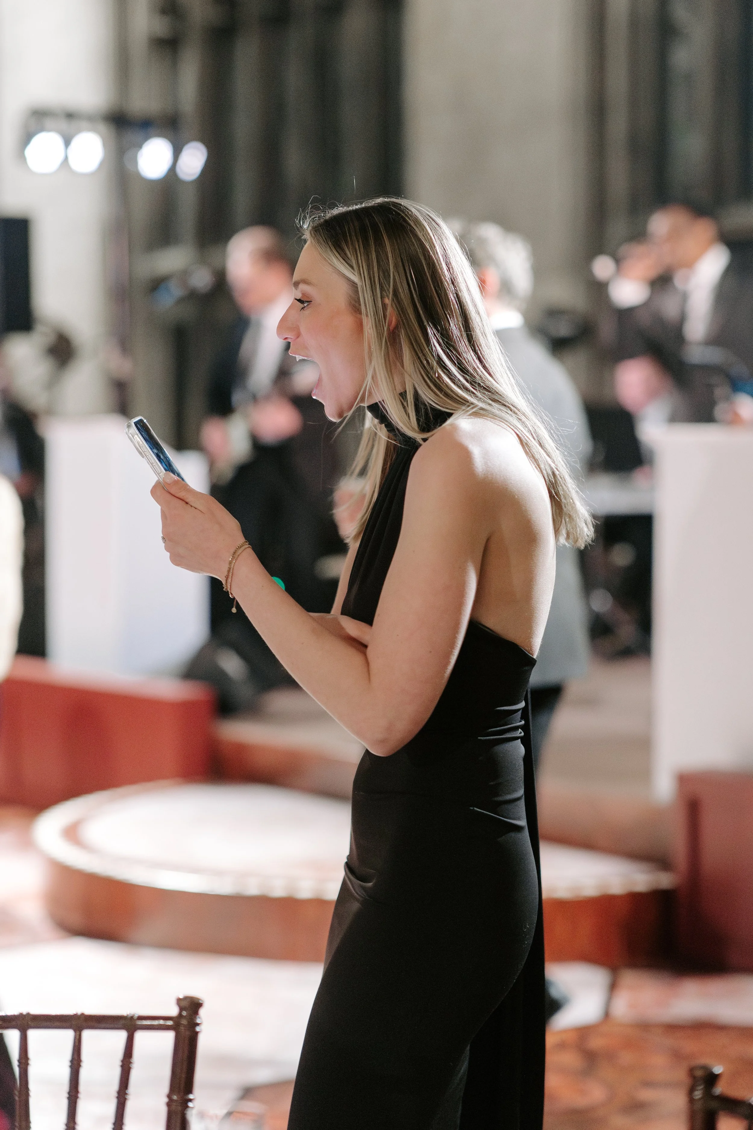 A woman in a black dress with blonde hair holding a smartphone, standing in a sophisticated setting with people in formal attire in the background.