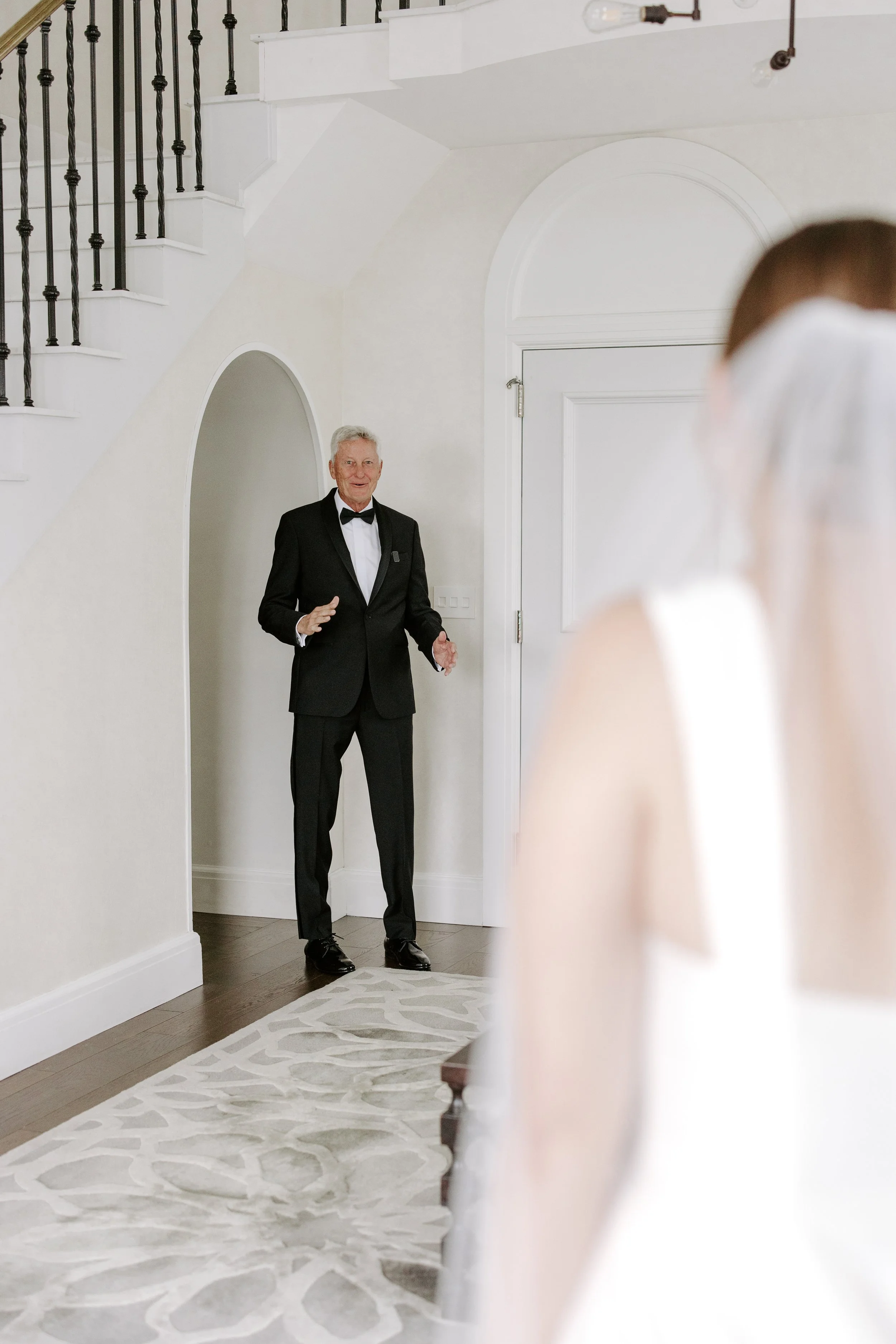 Older man in a tuxedo standing inside a white-walled room with stairs, talking to a woman with brown hair and a white veil, partially visible and blurred in the foreground.