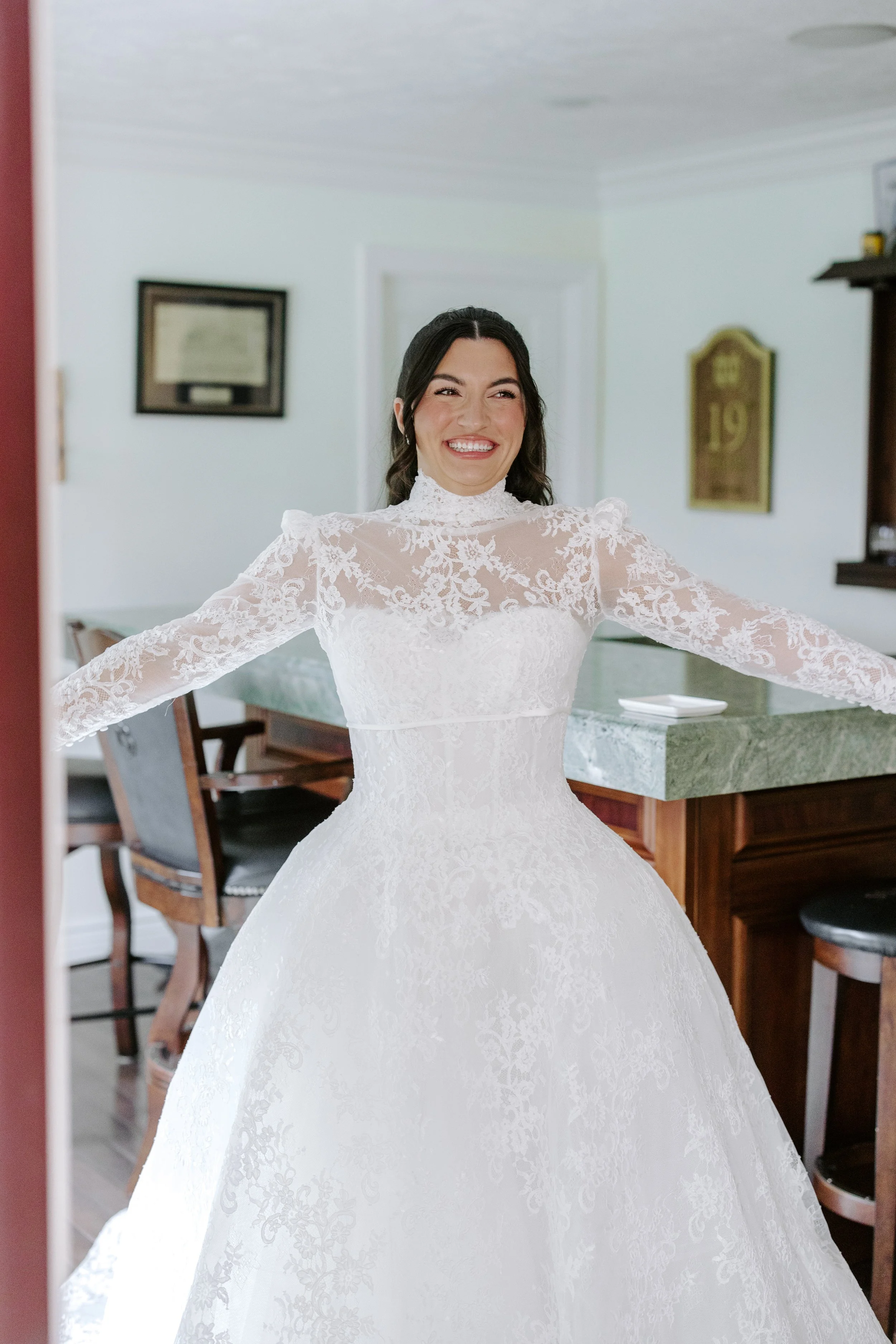 A woman in a white lace wedding dress smiling with her arms outstretched in a home interior.