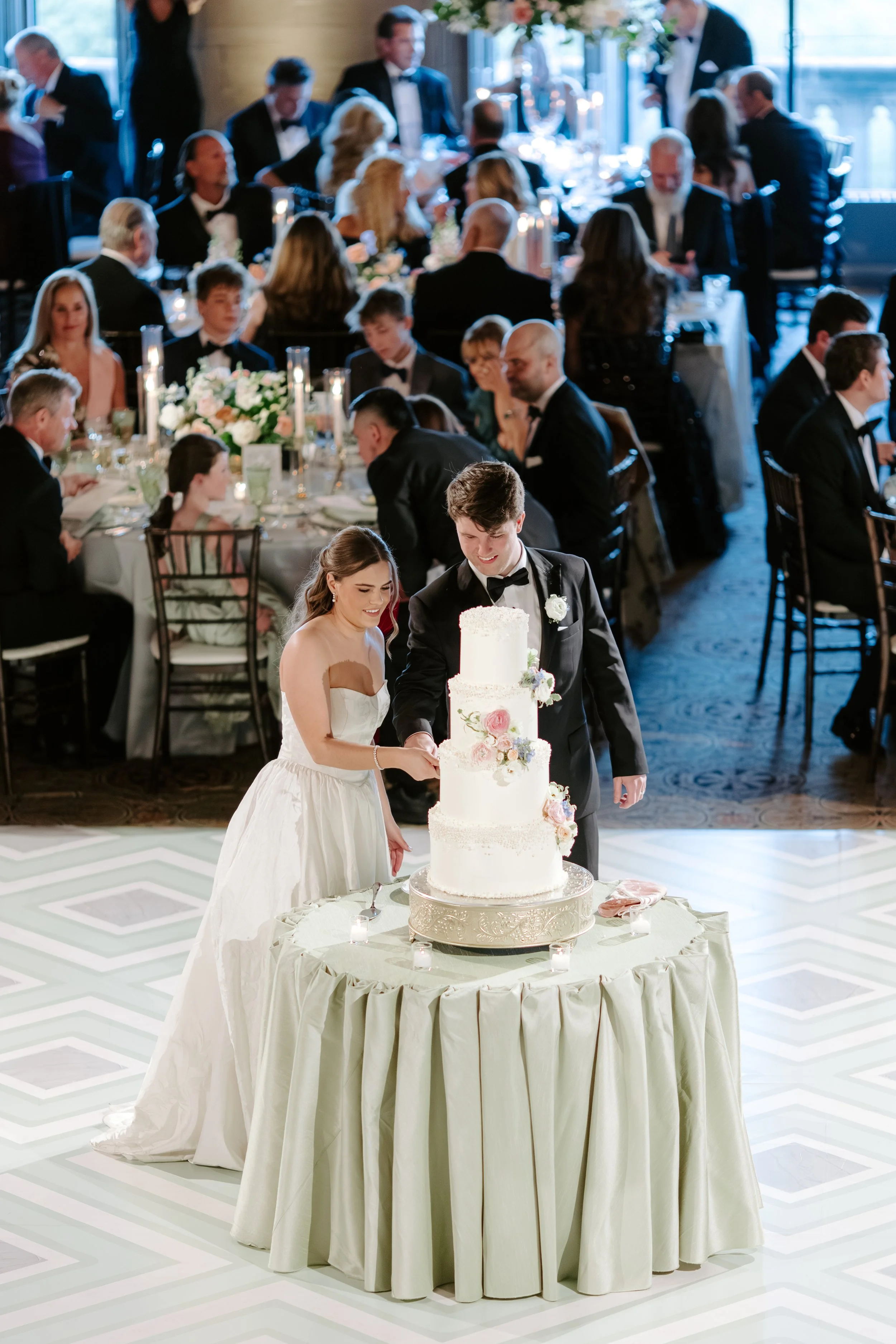 A bride and groom cutting a white multi-tiered wedding cake decorated with flowers at their wedding reception, with guests seated at tables in the background.