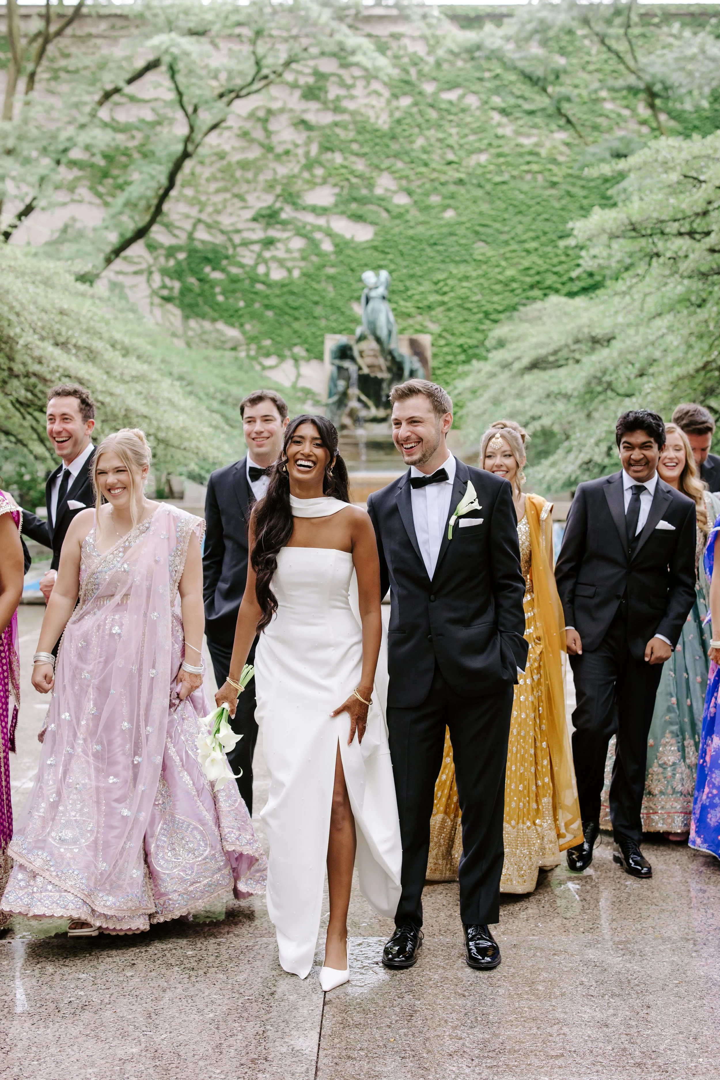 A diverse group of people in formal attire, including a bride in a white gown, walking outdoors on a paved path surrounded by lush green trees. In the background, there is a large fountain with a sculpture of a horse.