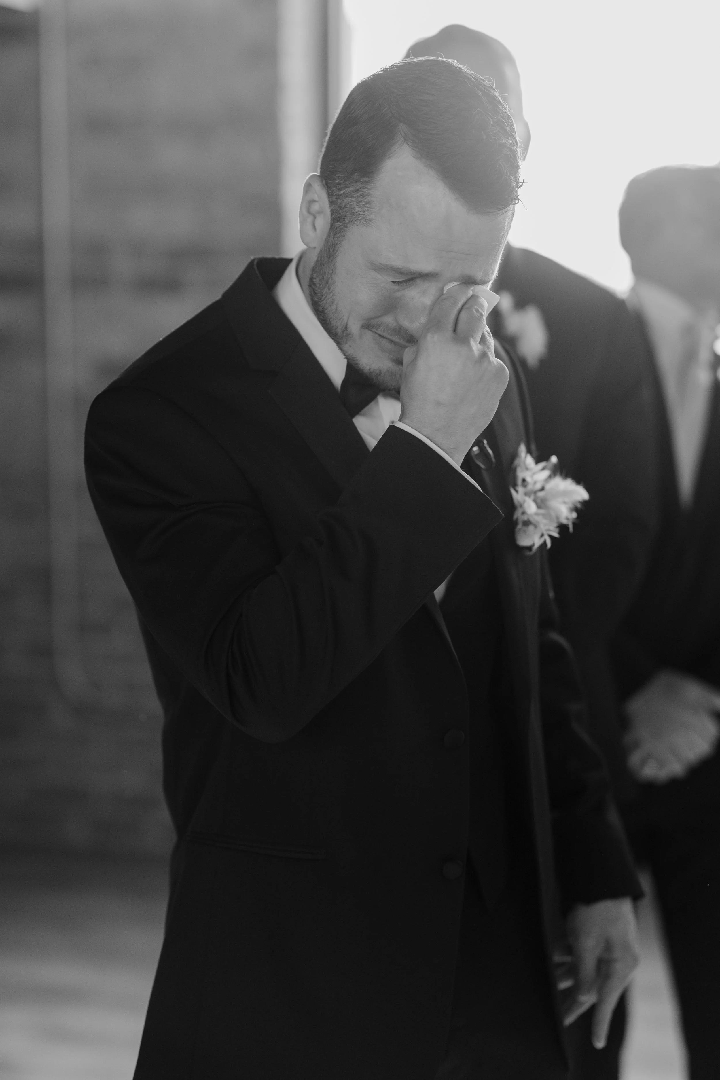 A man dressed in formal attire, possibly a tuxedo, is holding an object in his hand and appears to be experiencing an emotional moment, with his eyes closed and a hand covering part of his face. The background suggests a formal or special event setti