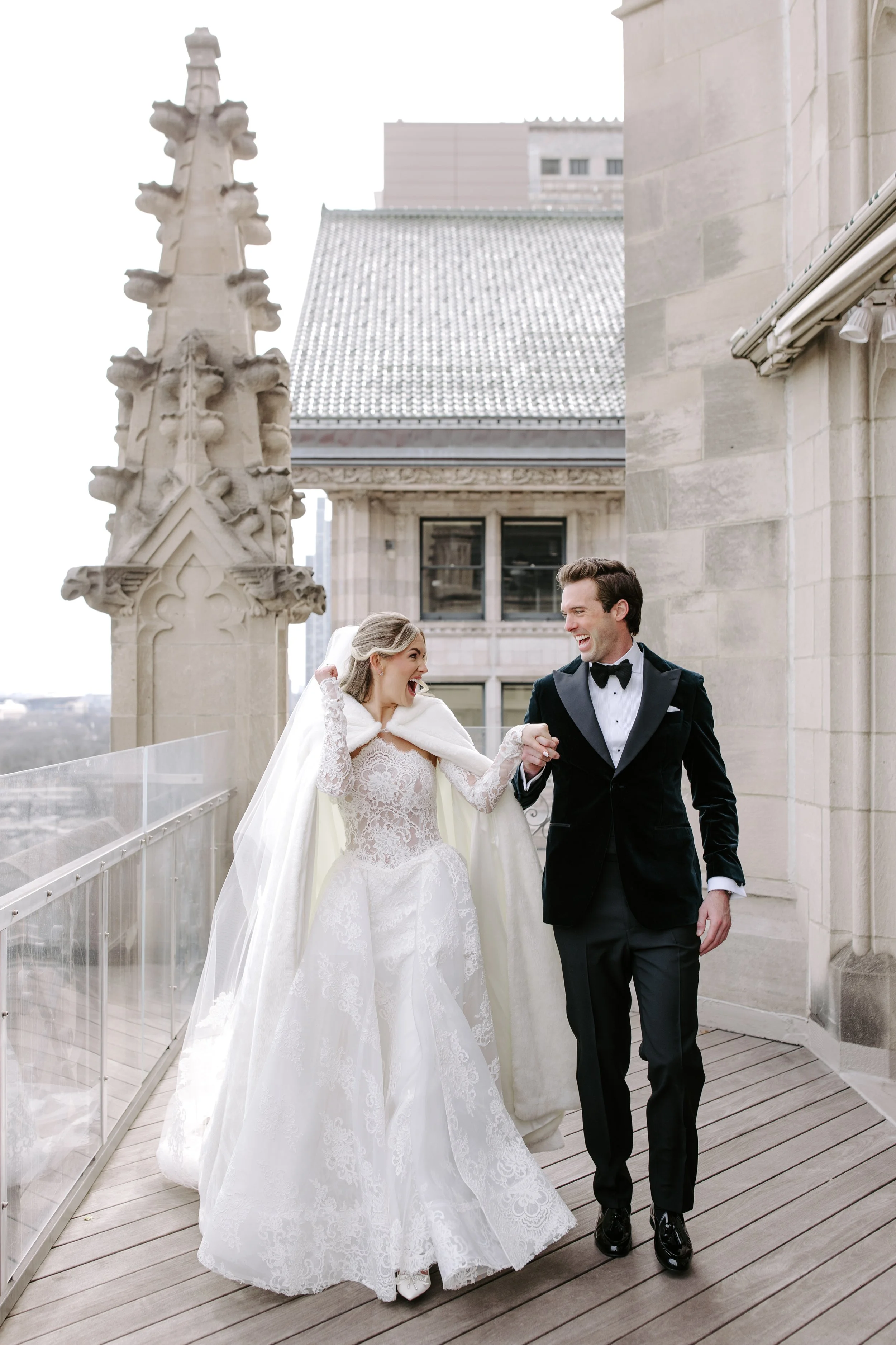 A bride and groom are holding hands and smiling on a rooftop terrace with historic architecture in the background.