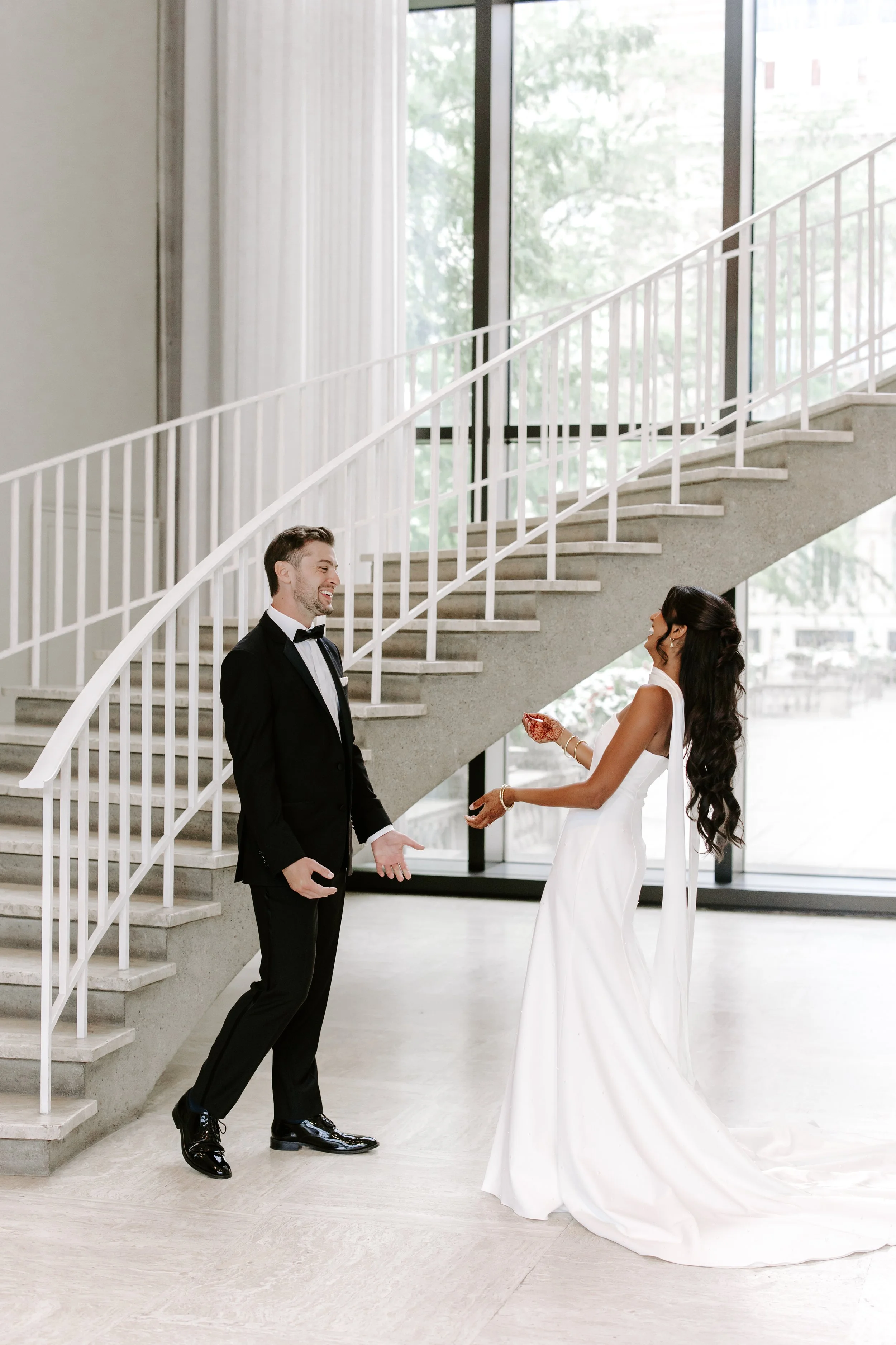 A happy bride and groom at their wedding, standing indoors near a staircase with large glass windows in the background, exchanging vows.