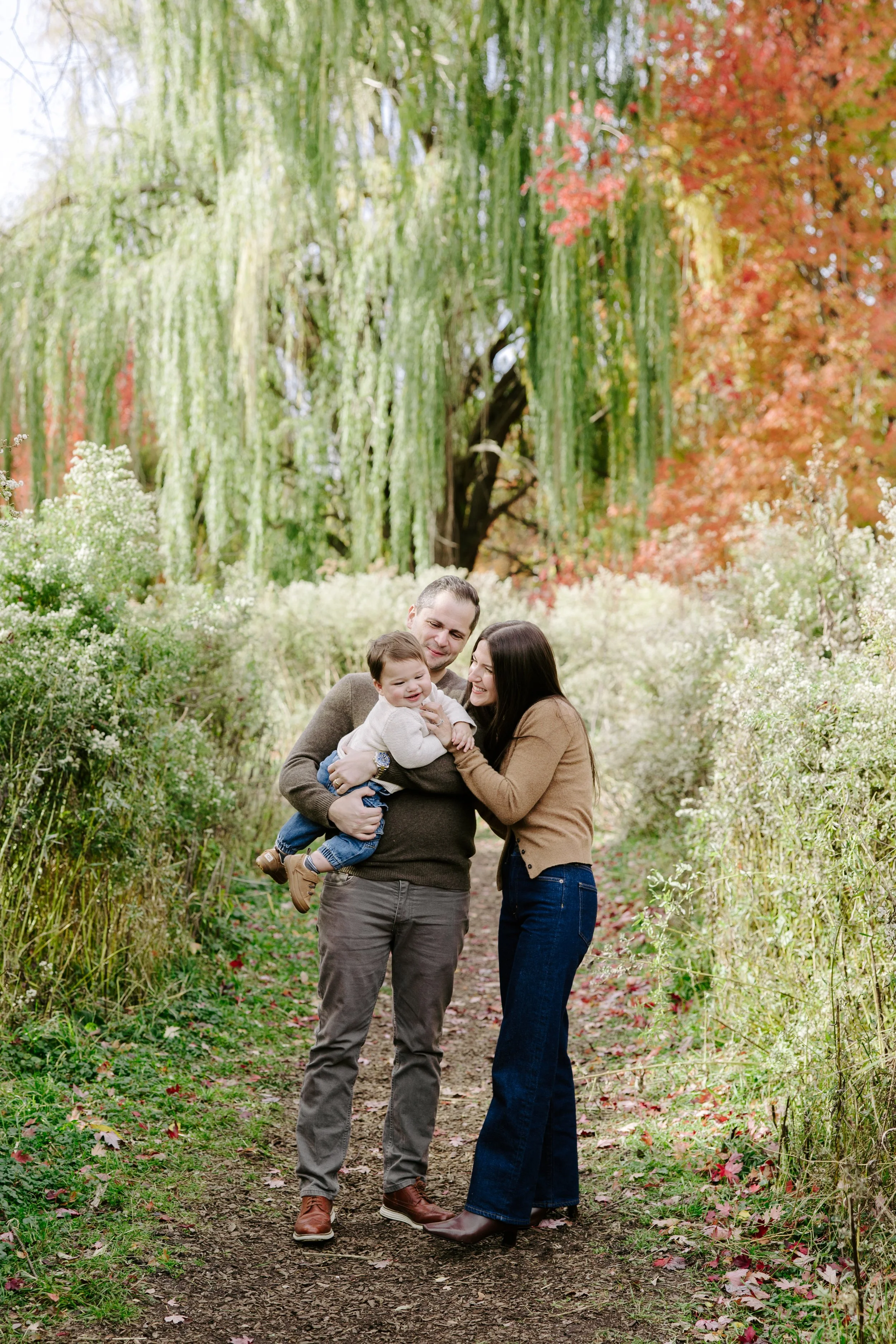 A family of three, a man, woman, and a young child, standing on a dirt path in a park with lush greenery and fall foliage. They are smiling and enjoying each other's company.