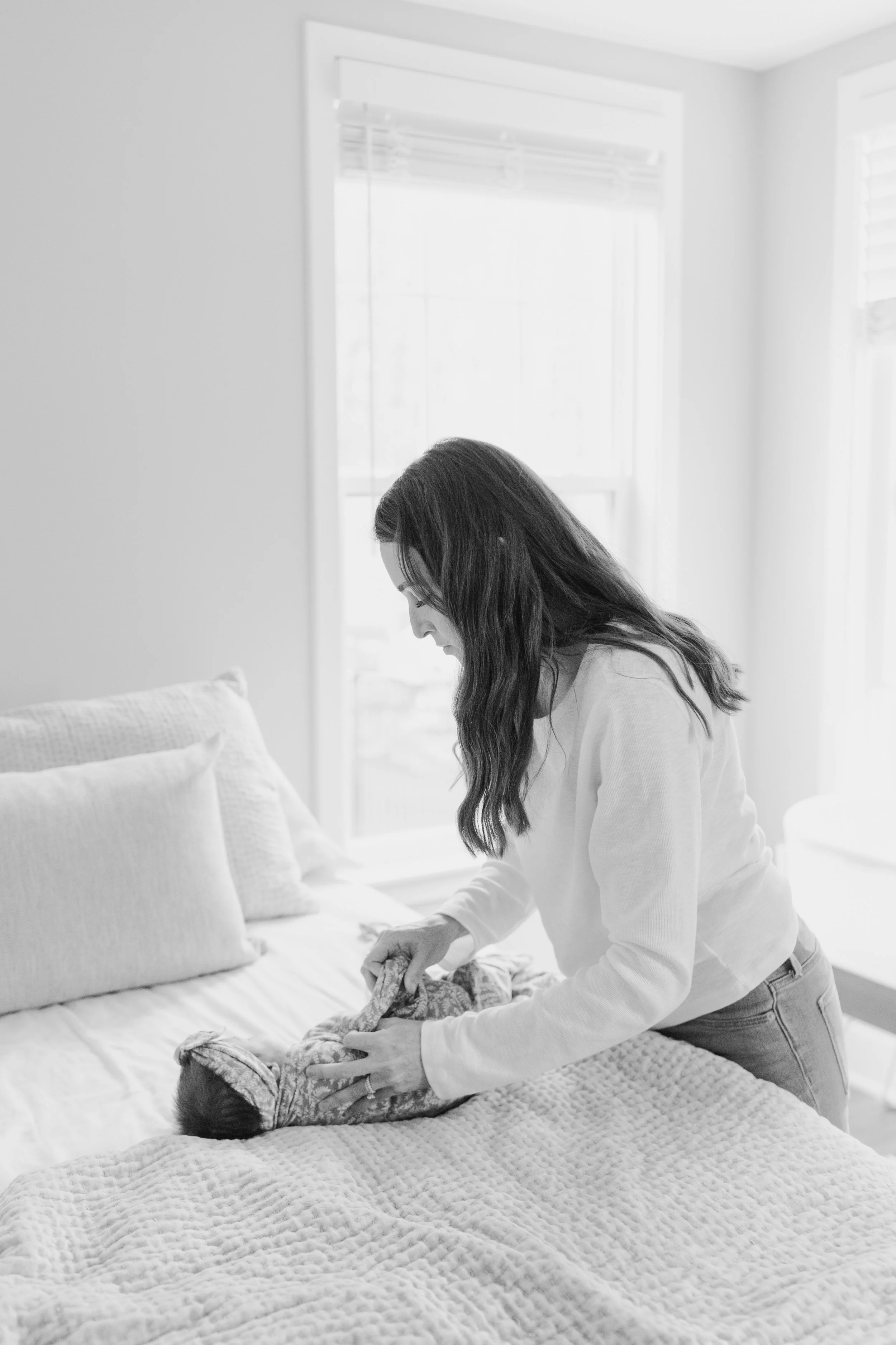 A woman with long dark hair standing by a bed with white bedding, looking down at a person lying in bed with a blanket over their head, in a bright room with large windows.