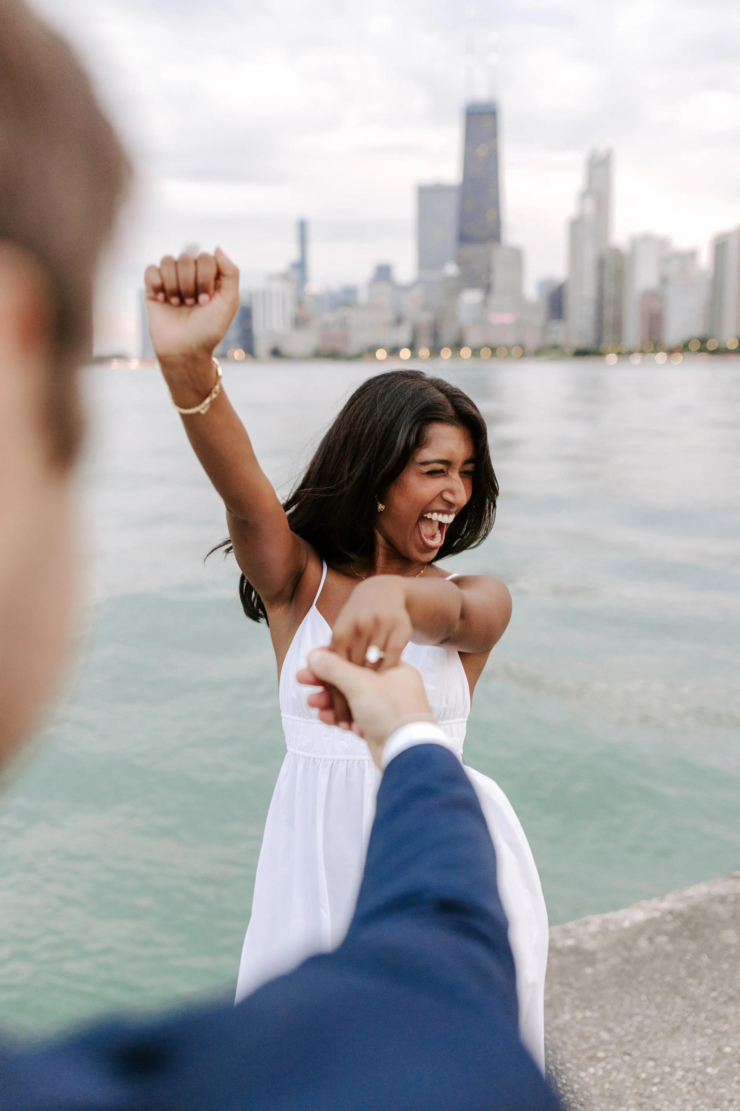 A woman in a white dress standing near water, smiling and raising her arm, with city skyscrapers in the background.