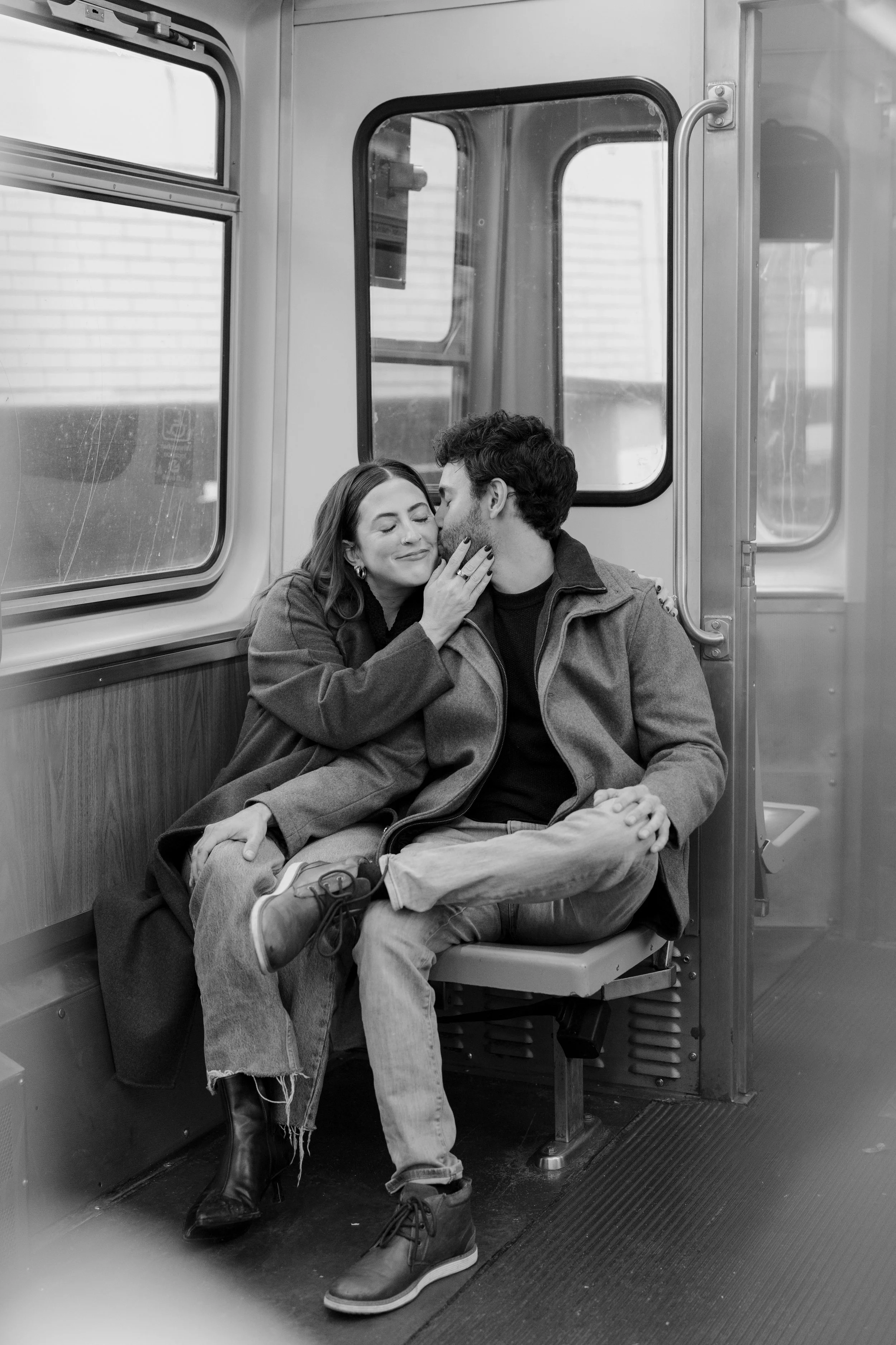 A couple sitting close together in a subway train, sharing a tender moment with one person kissing the other on the cheek, both smiling peacefully.