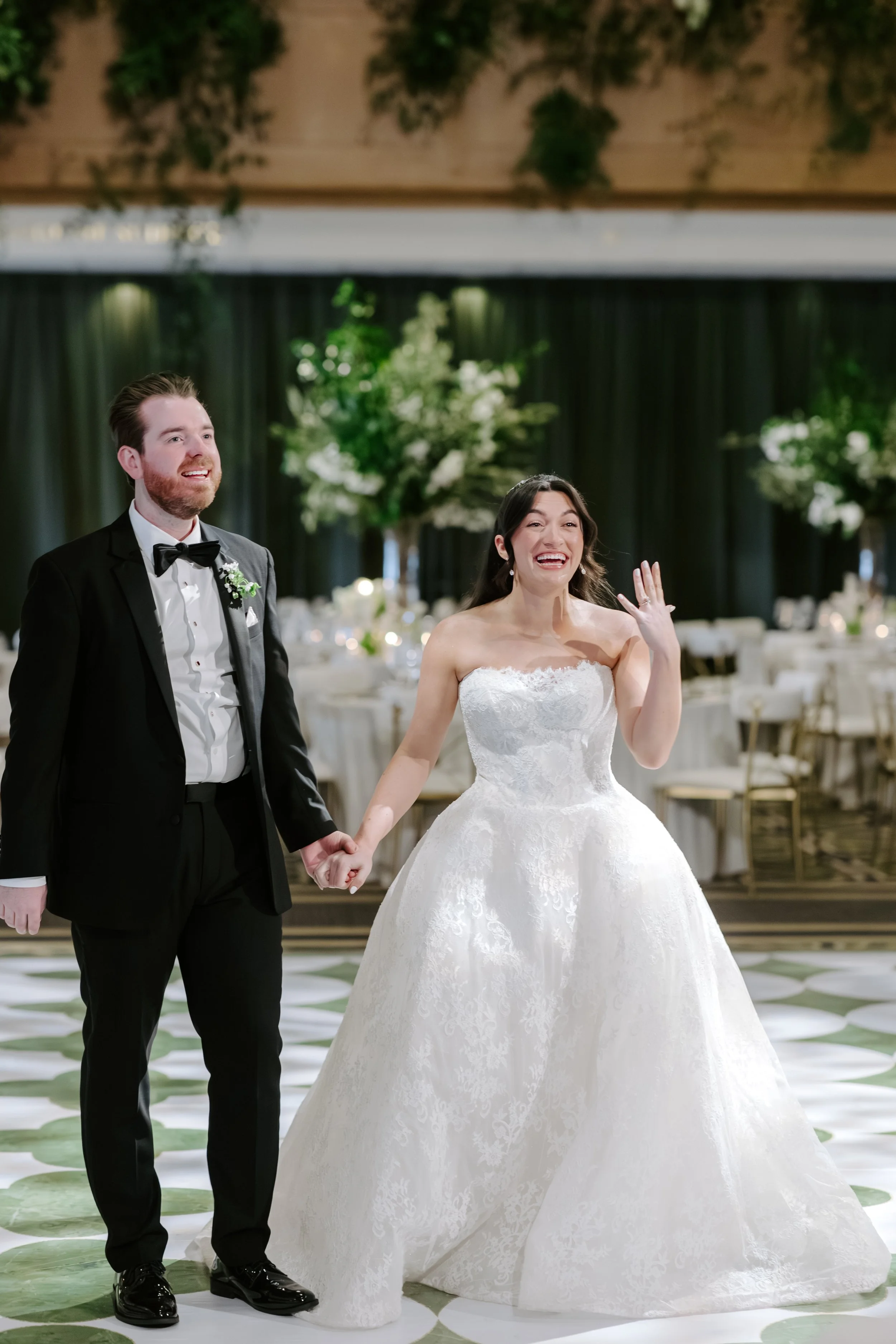 A bride and groom hold hands at their wedding reception, smiling and looking happy. The bride wears a white wedding gown and the groom wears a black tuxedo with a bow tie. The setting is decorated with floral arrangements and elegant tables in the ba