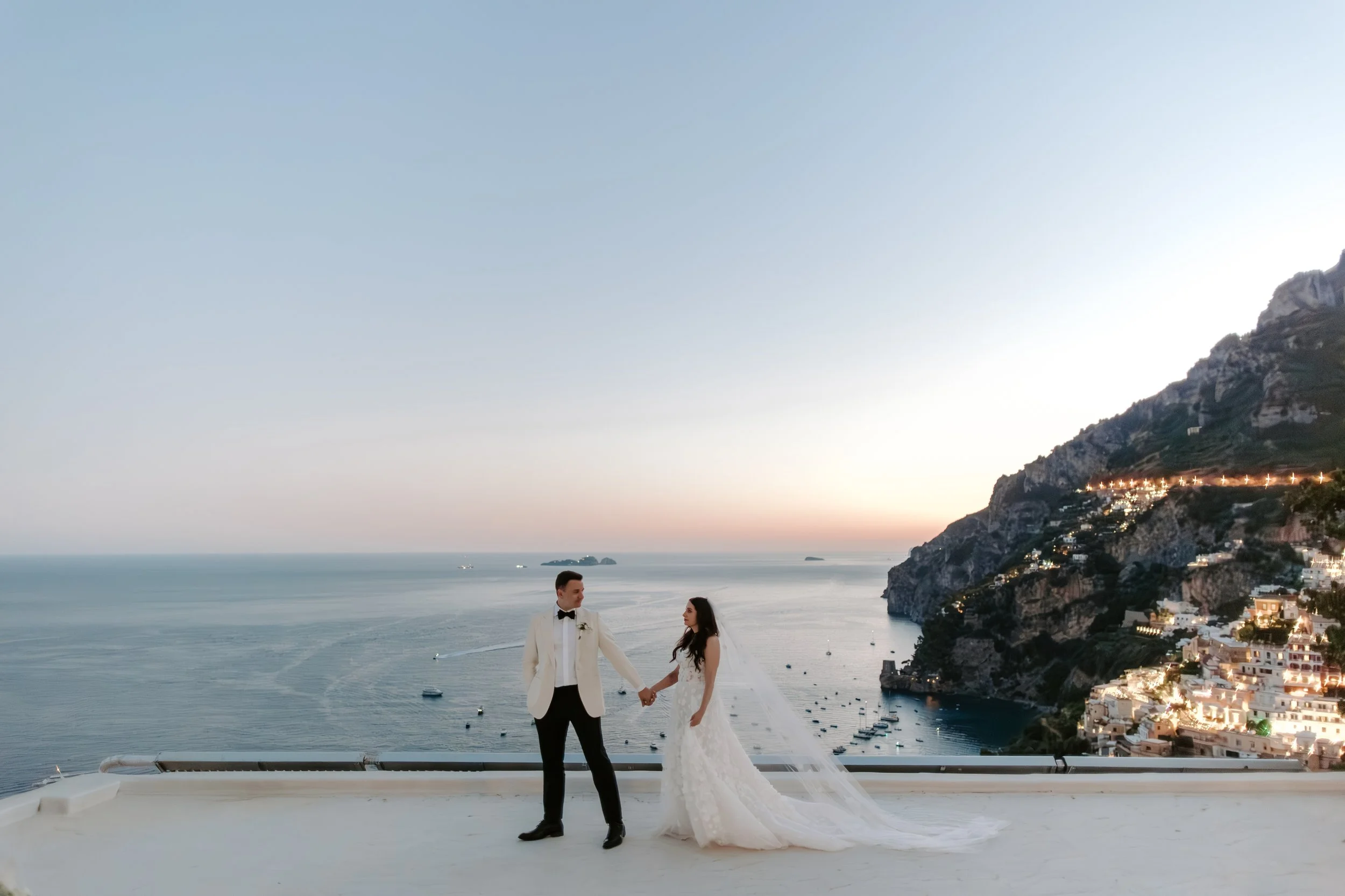 A couple dressed in wedding attire holding hands on a white platform overlooking a seaside town with cliffs and buildings illuminated at dusk.