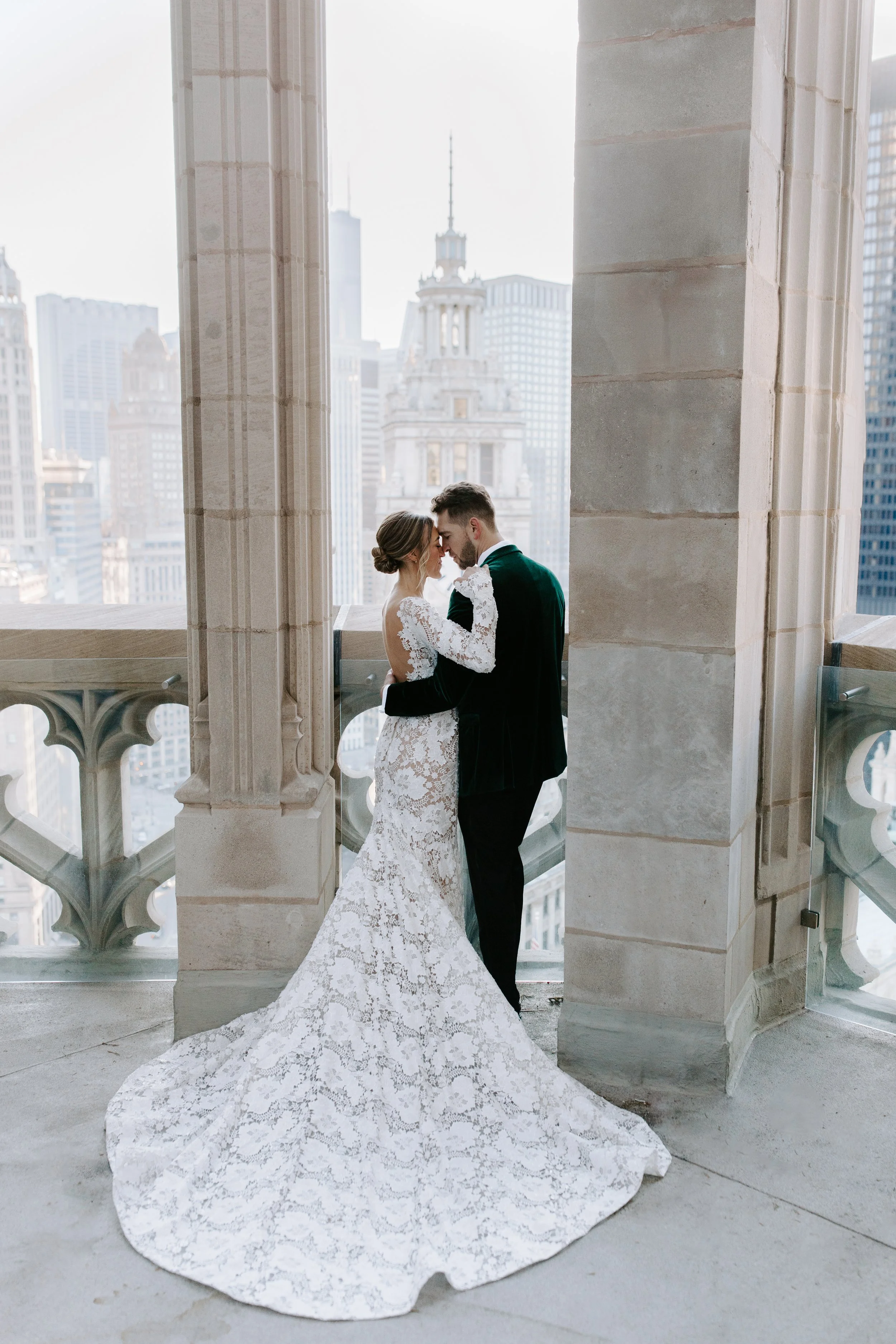 A newlywed couple sharing a romantic moment on a balcony overlooking a city skyline with tall buildings and a historic clock tower.