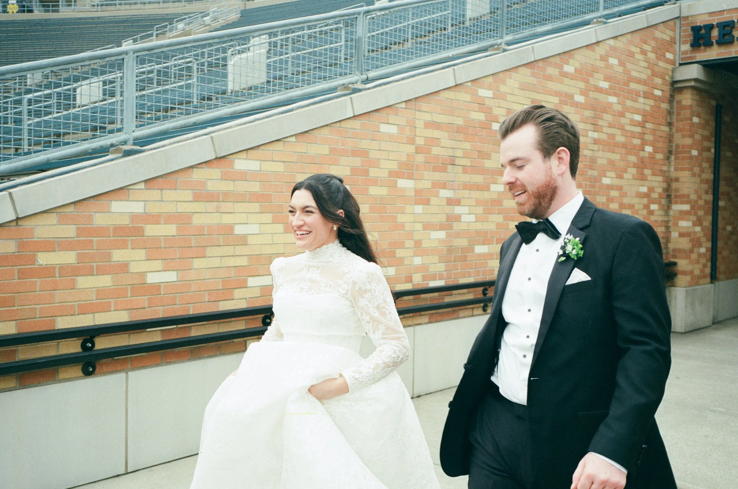 A newly married couple walking outdoors in front of a brick building; the bride wears a white lace wedding dress, and the groom wears a black tuxedo with a bow tie and boutonniere.