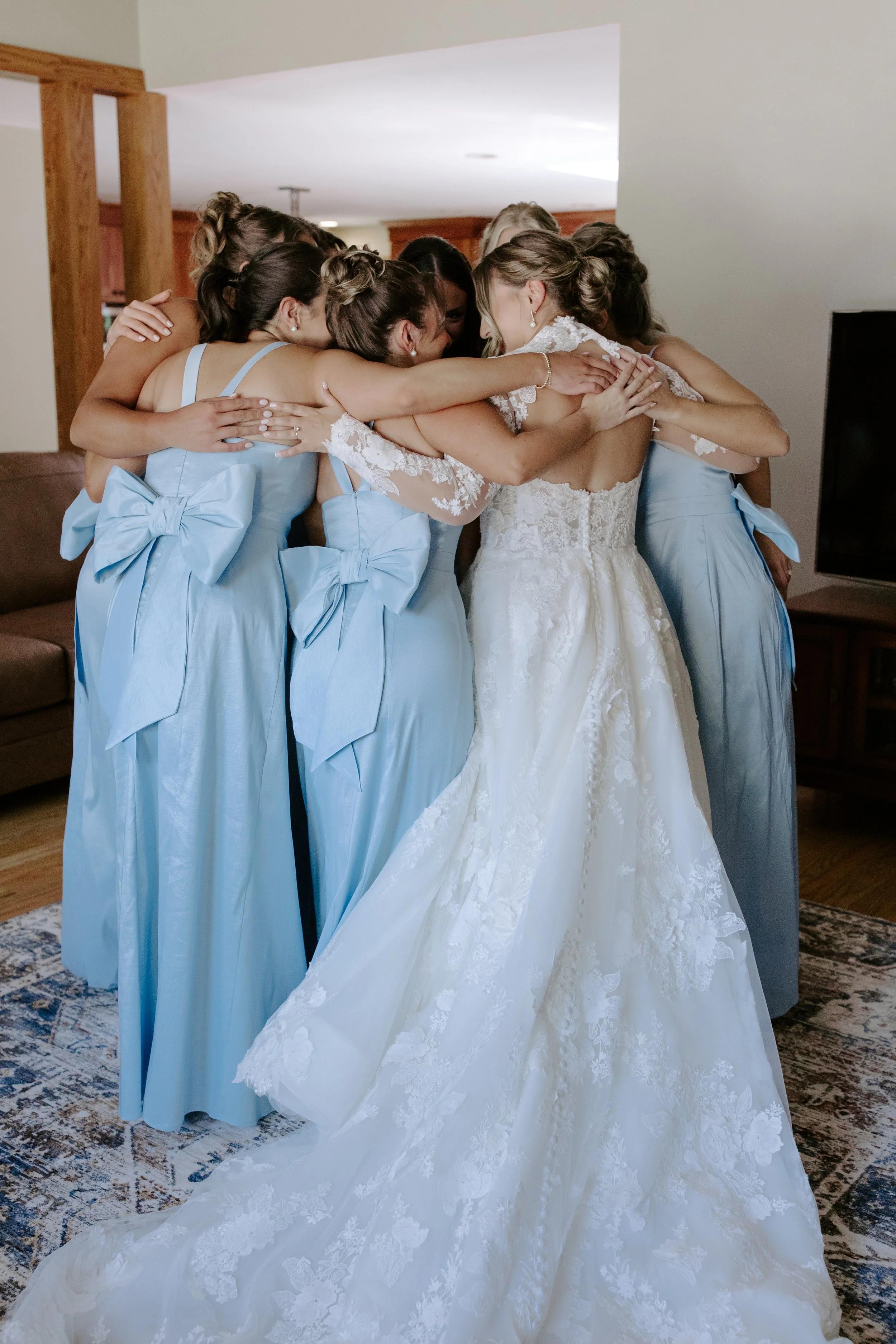 Bride in a white lace wedding gown huging bridesmaids in blue dresses.
