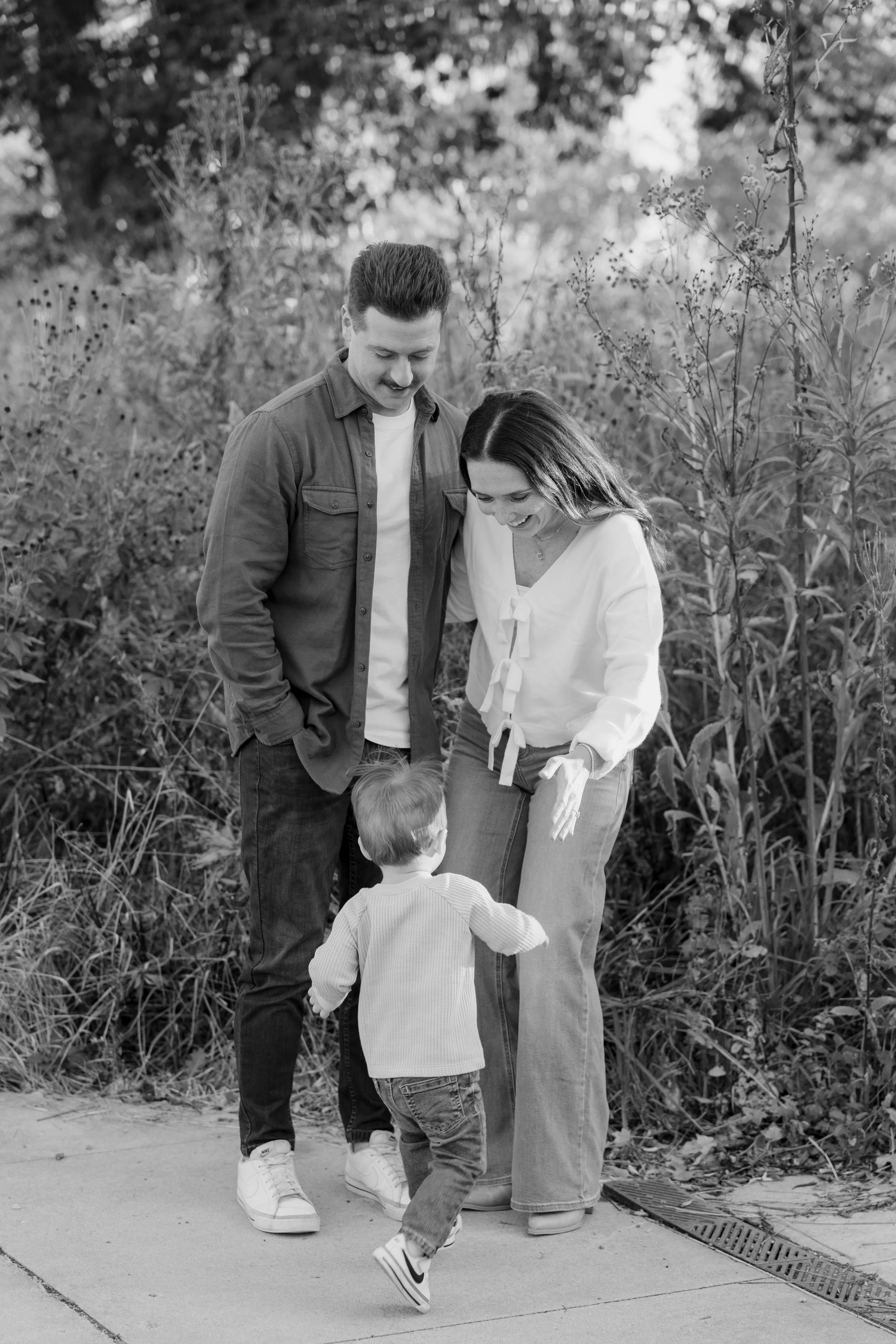 A family of four walking outdoors on a sidewalk, with a man, woman, and two children, surrounded by trees and foliage.