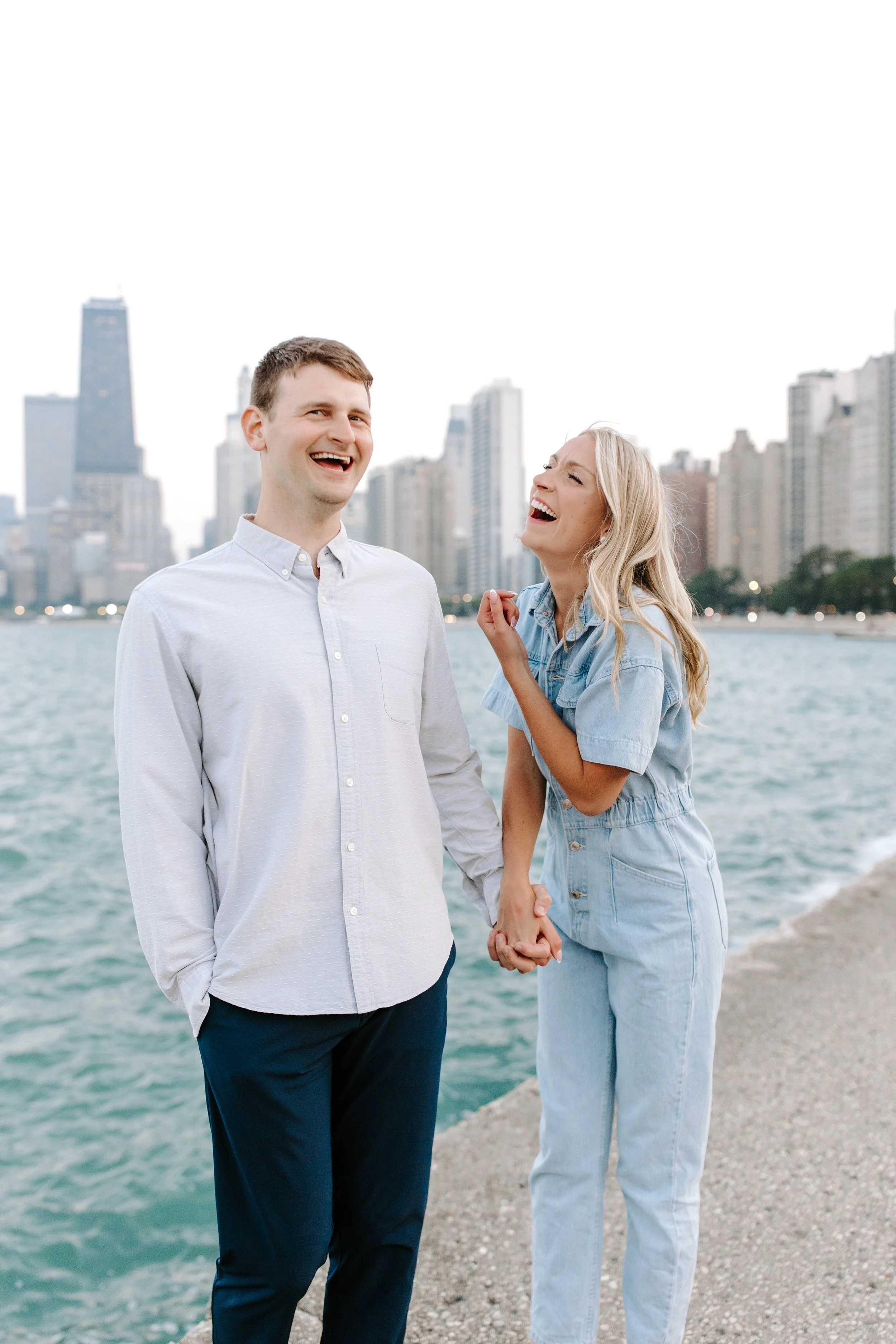 A smiling man and woman holding hands near a body of water with city skyscrapers in the background.