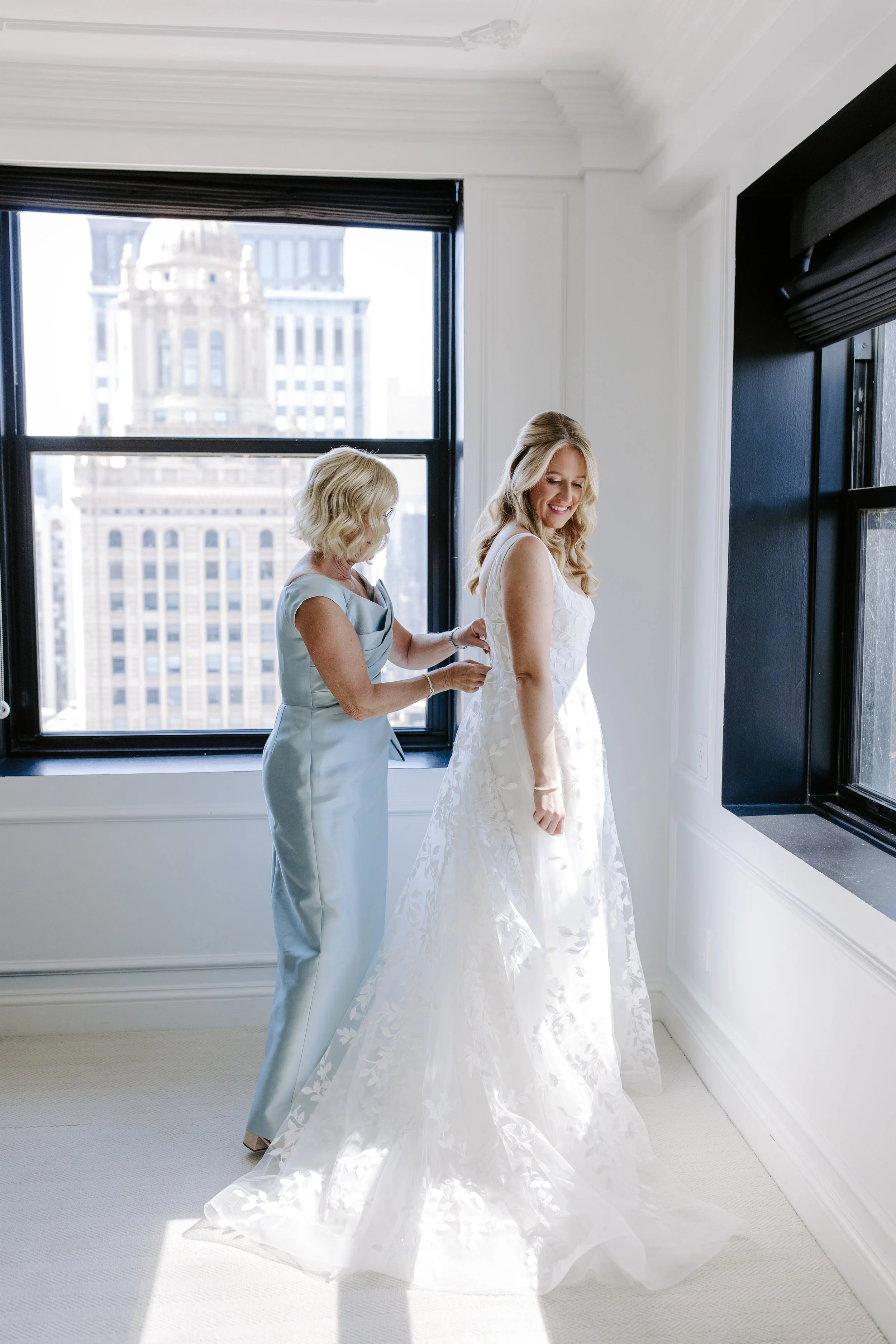 A bride in a white wedding dress smiling as an older woman, possibly her mother, helps her with her dress in a bright room with tall windows showing city buildings outside.