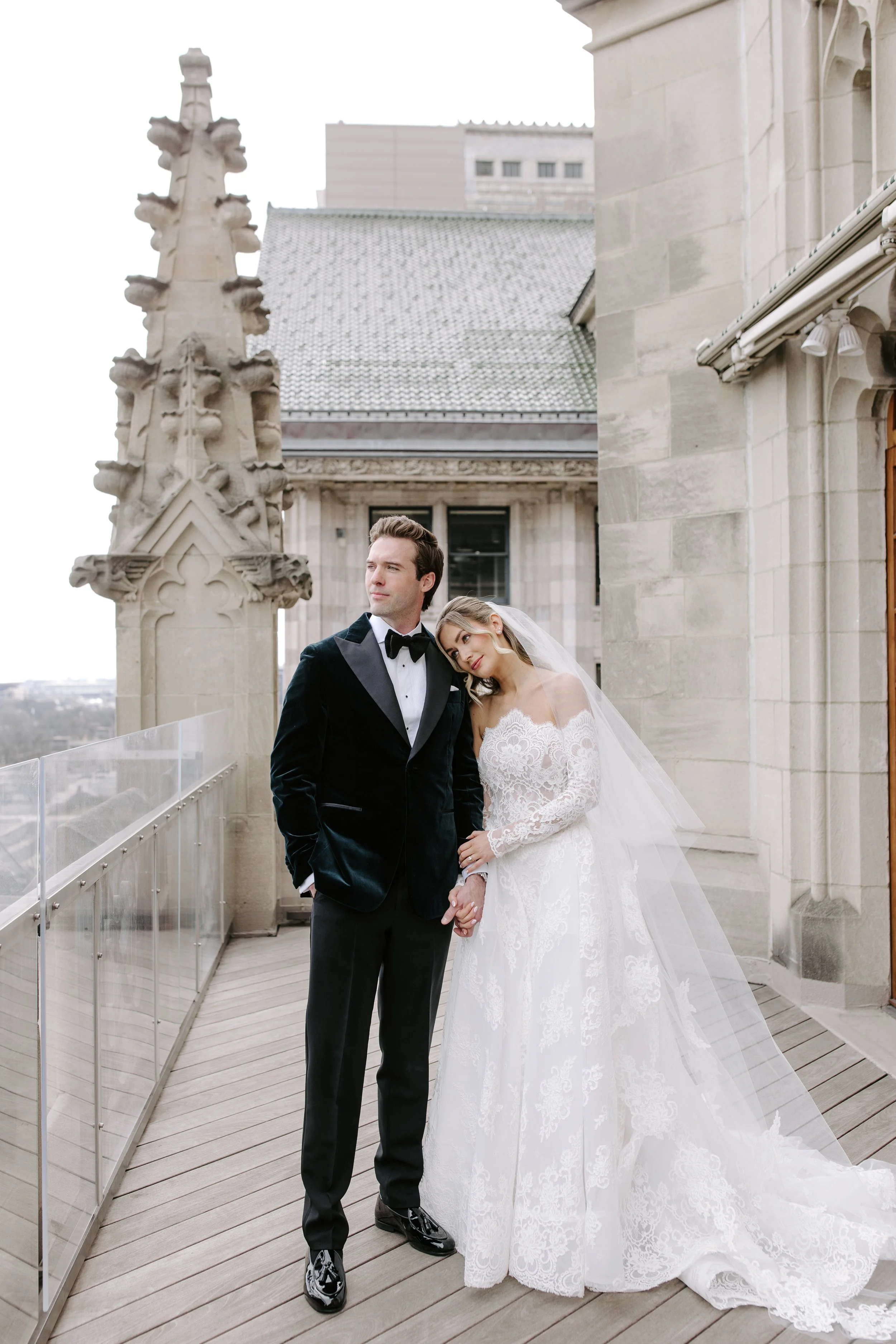 A bride and groom standing on a rooftop terrace with architectural details and city skyline in the background. The bride is leaning her head on the groom's shoulder. They are dressed in wedding attire; the groom in a black tuxedo and the bride in a w