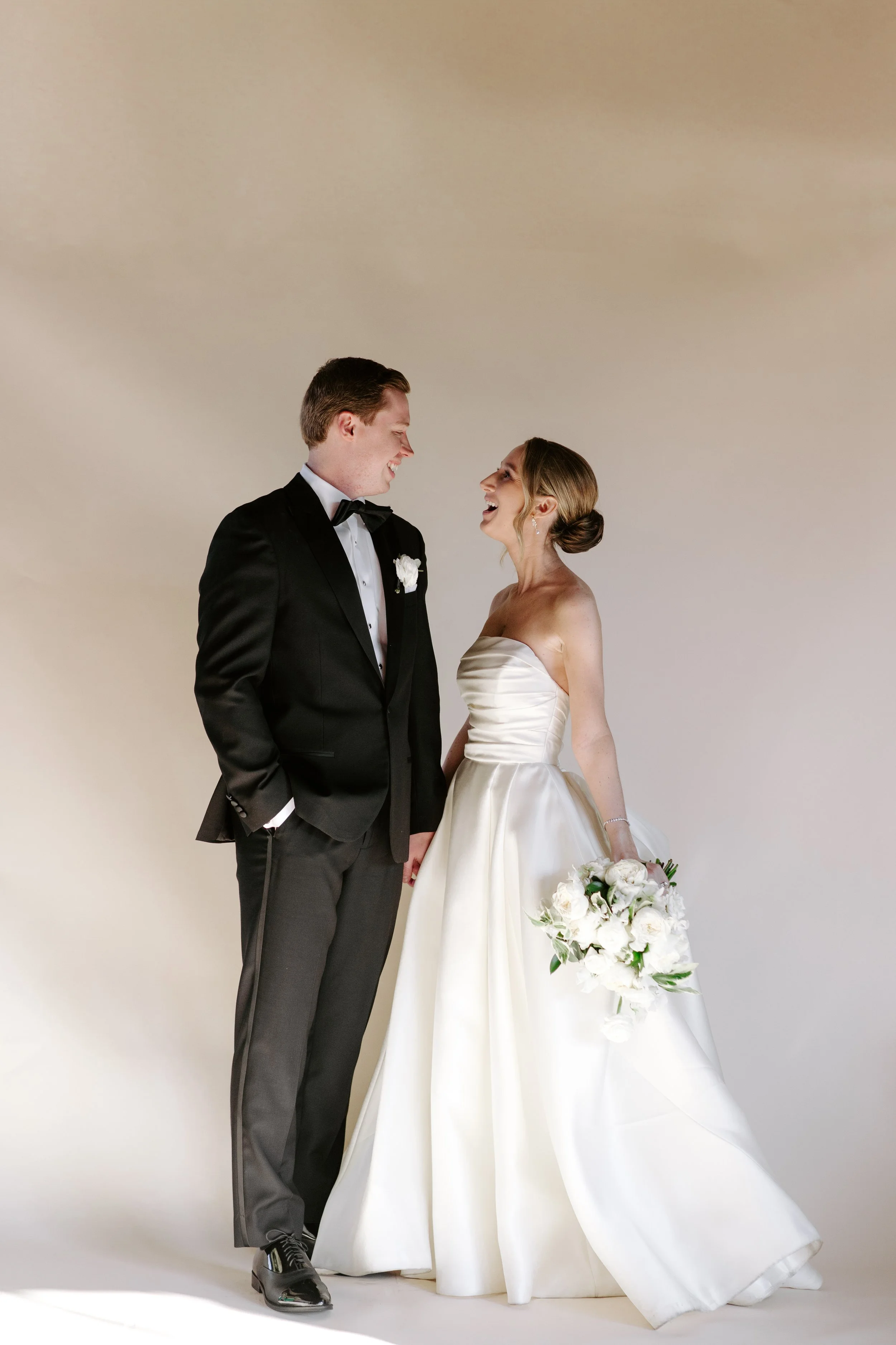 A bride and groom are smiling at each other and holding hands, standing against a plain background. The groom is wearing a black tuxedo with a bow tie, and the bride is in a strapless white wedding gown, holding a bouquet of white flowers.