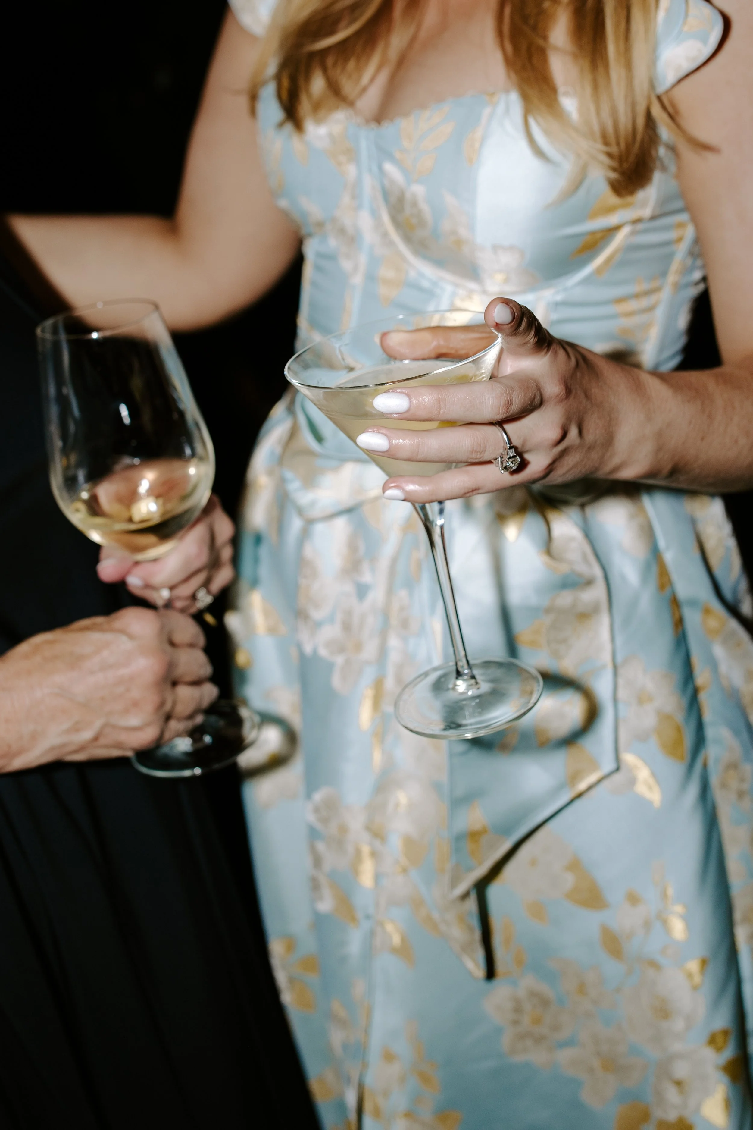 A woman in a blue and gold floral dress holding a martini glass with a clear beverage, and another person holding a wine glass with white wine. The woman's face is not visible.