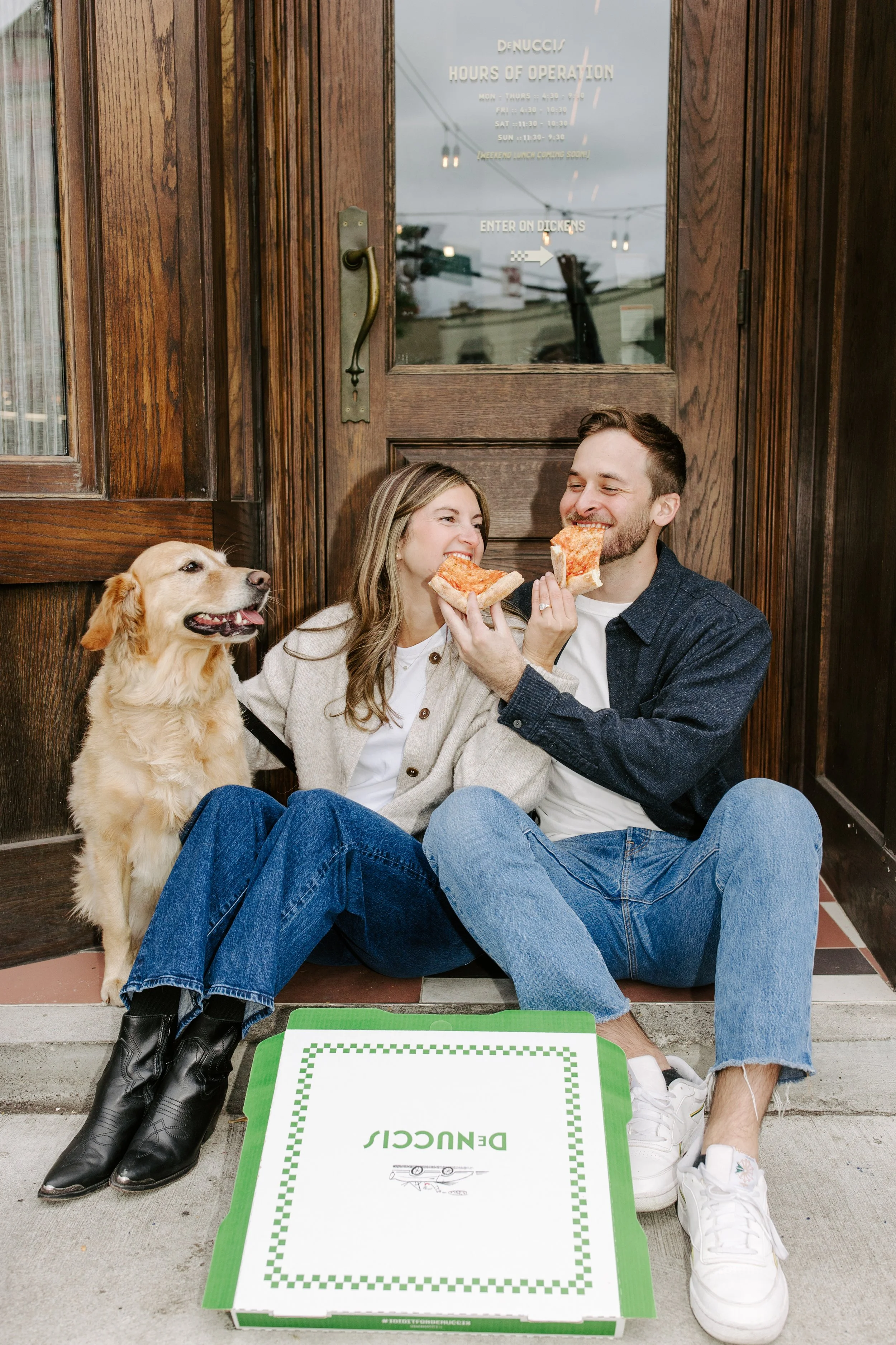 A young couple and a dog sitting outside a restaurant with a pizza box and a green and white pizza box on the sidewalk. The couple is eating pizza and smiling at each other, while the dog looks on. The restaurant has wooden doors with a sign listing 