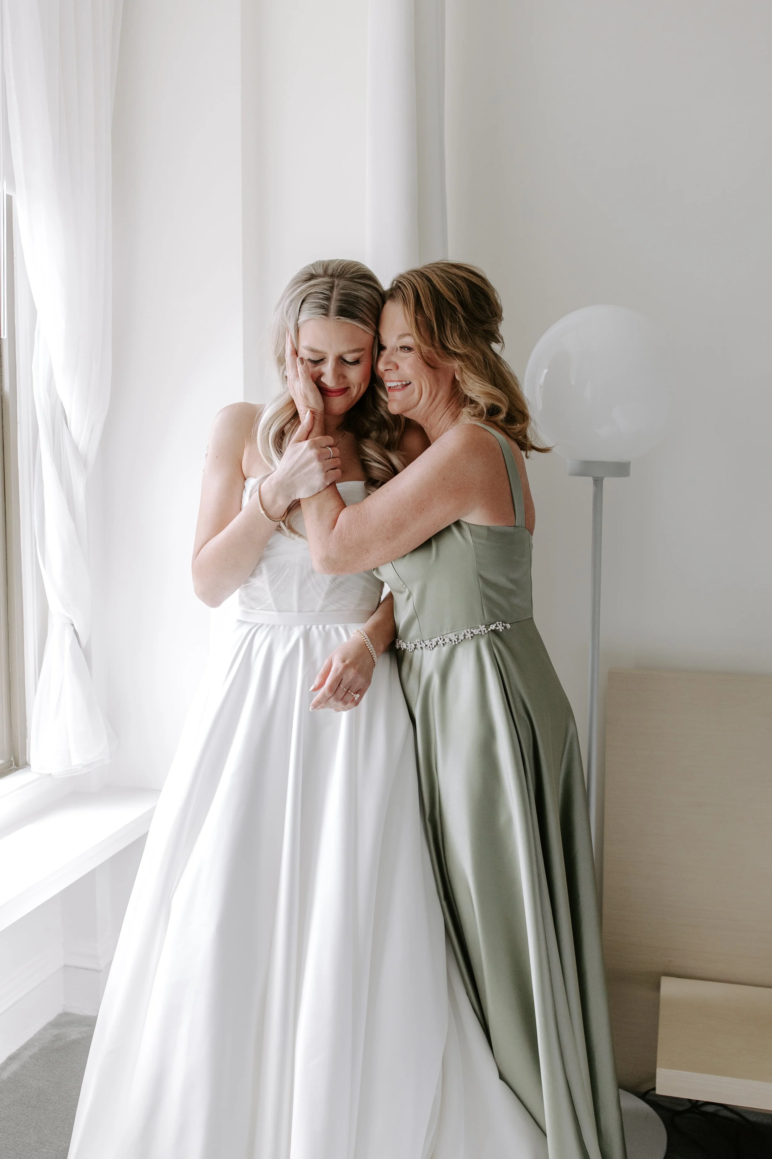 A bride and a woman, possibly her mother or a close relative, sharing an emotional moment in a bright, minimalistic room. The bride has long blonde hair and is wearing a white wedding gown. The other woman has shoulder-length wavy hair and is wearing