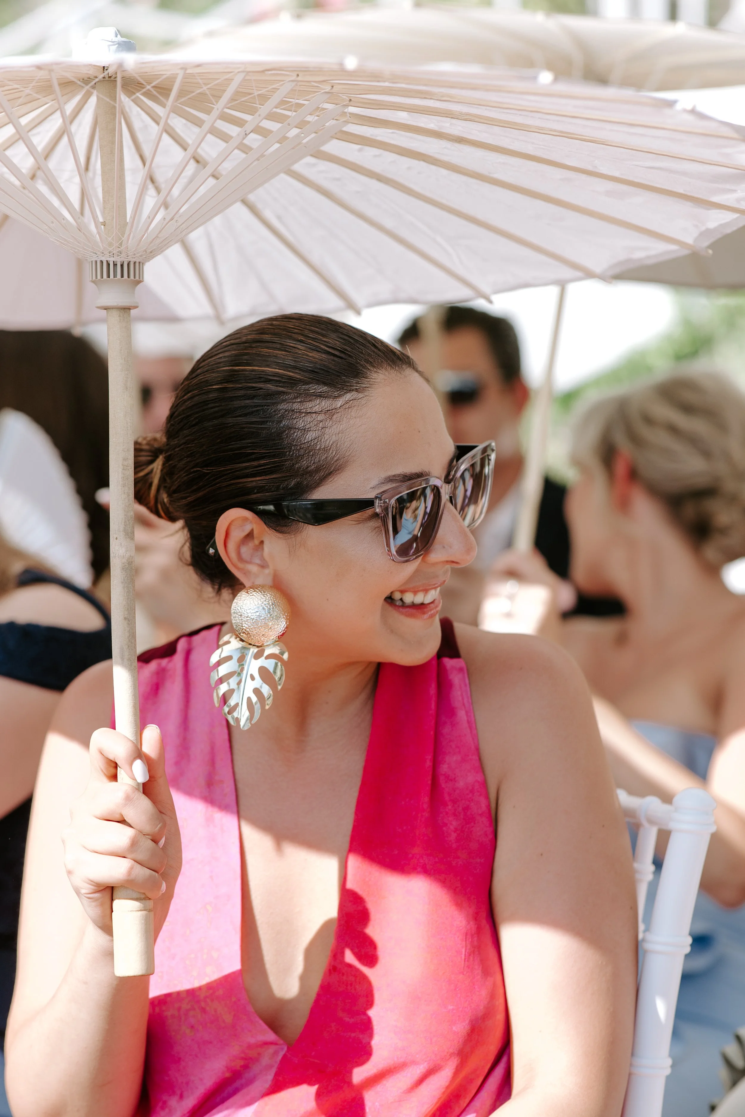 A woman wearing sunglasses and large earrings, holding a white parasol, smiling outdoors on a sunny day.
