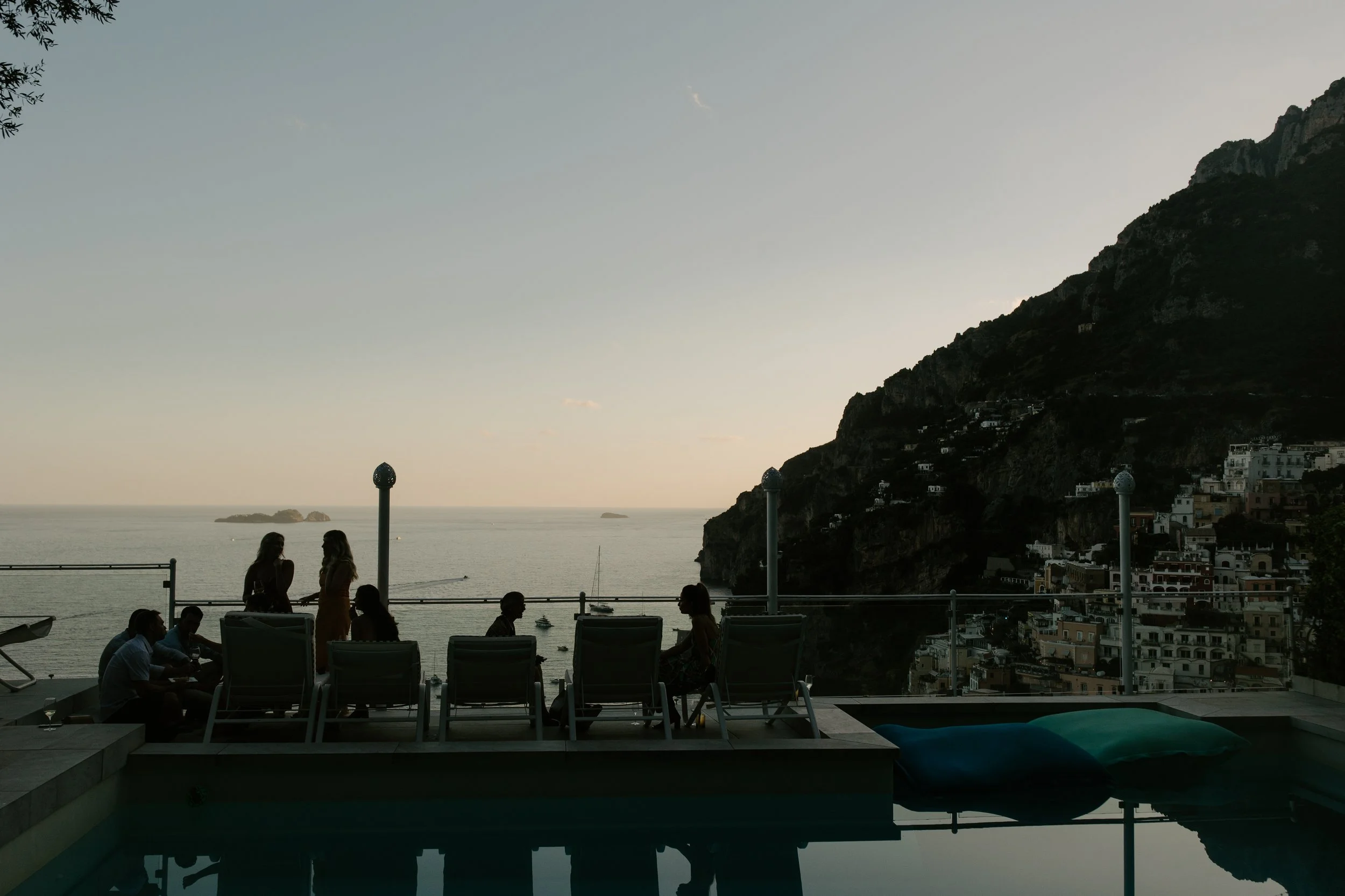 People seated outdoors at a restaurant with ocean and hillside town in the background at dusk.