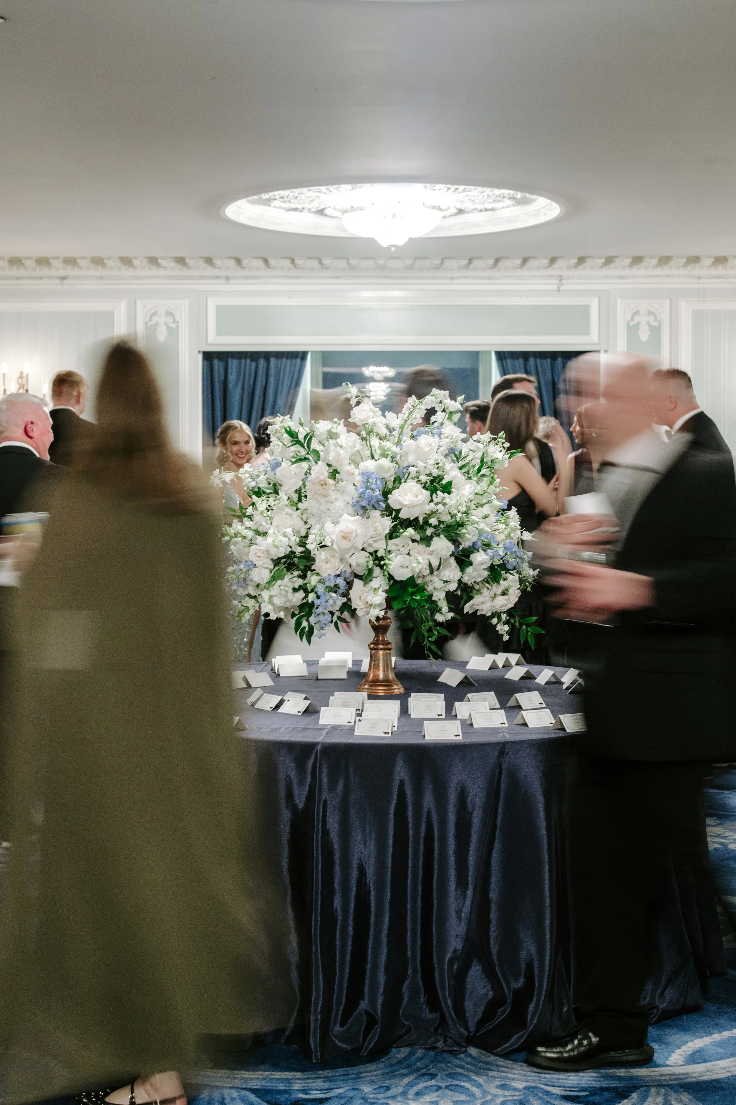 A formal event with people dressed in tuxedos and evening gowns, gathered around a table with a large floral centerpiece of white and light purple flowers, and name cards on a dark tablecloth, in a decorated banquet room with ornate ceiling details a