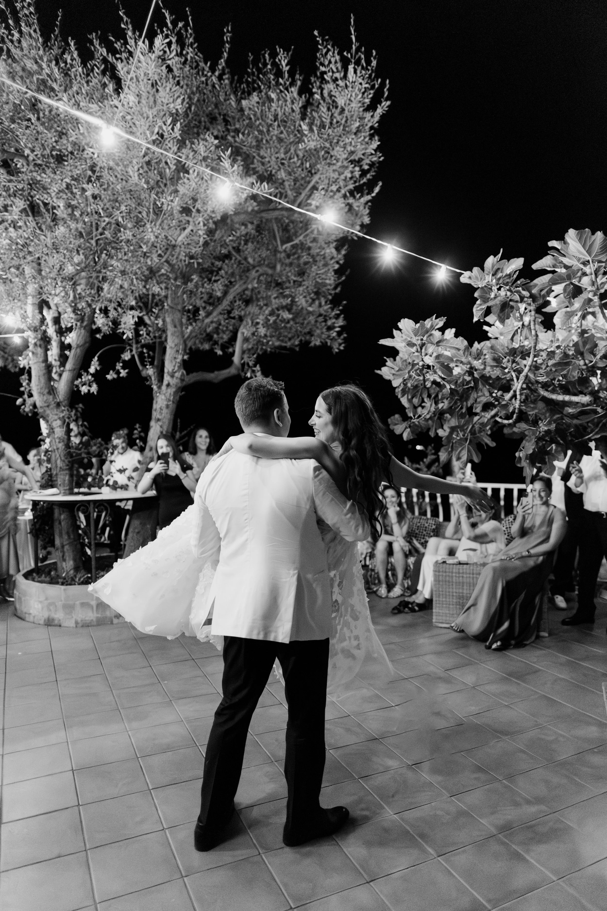 A bride and groom dancing outdoors at night under string lights, surrounded by guests watching and taking photos, with trees and a tiled floor in the background.
