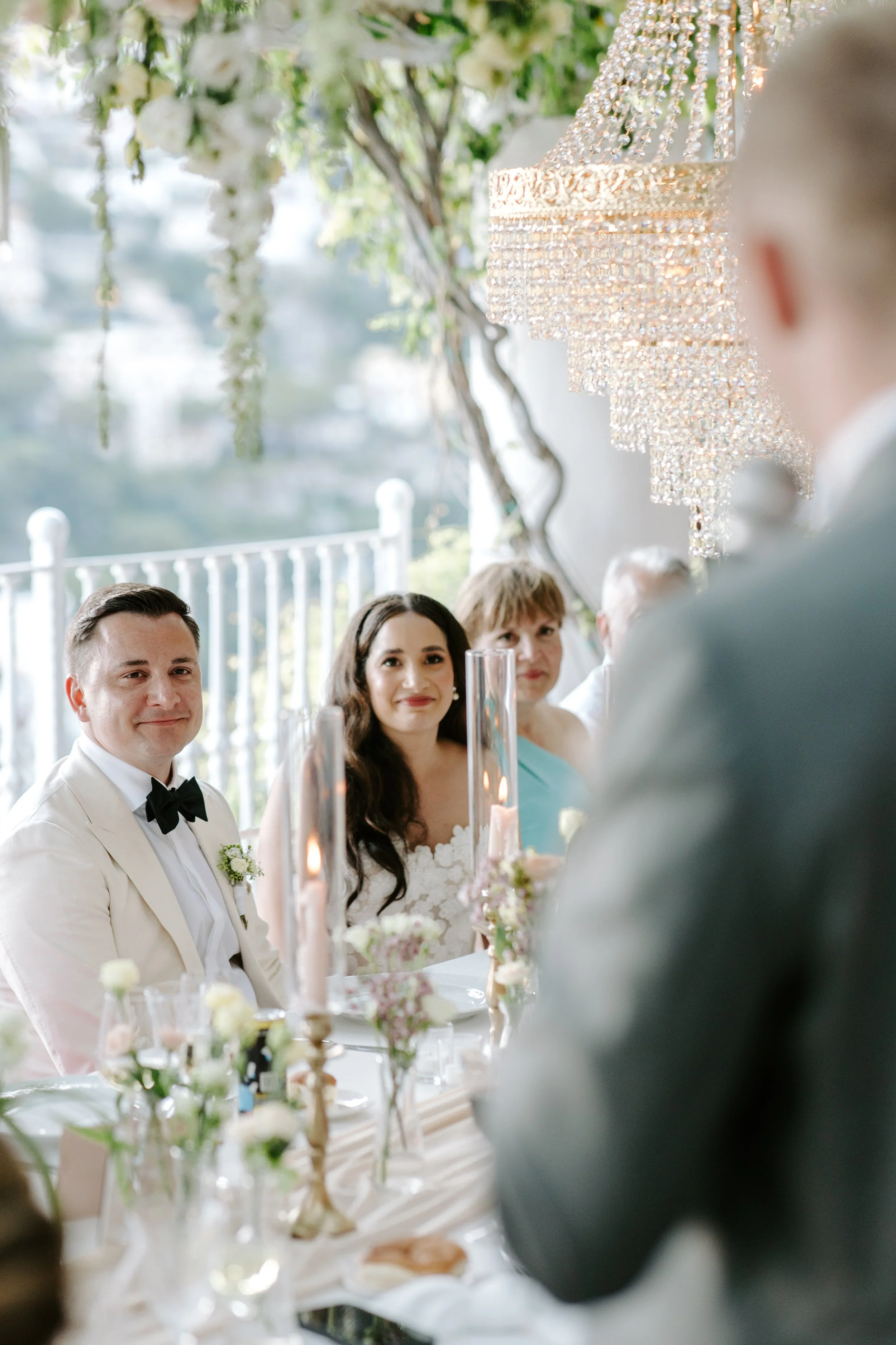 Guests sitting at a wedding reception table, with a focus on a man in a white tuxedo, woman with long dark hair, and an older woman, under a ceiling decorated with a large crystal chandelier and hanging floral arrangements.