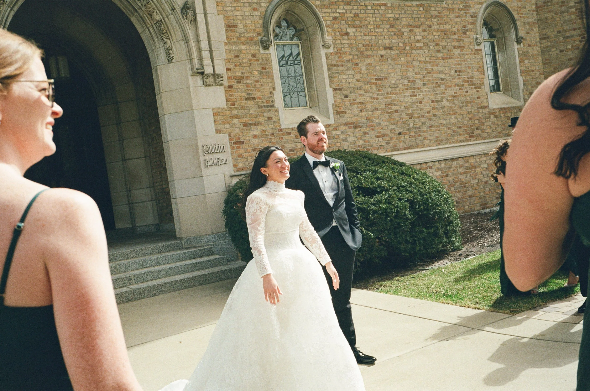 A bride and groom standing outside a church, smiling, with three women in dresses and one person with glasses in the foreground.
