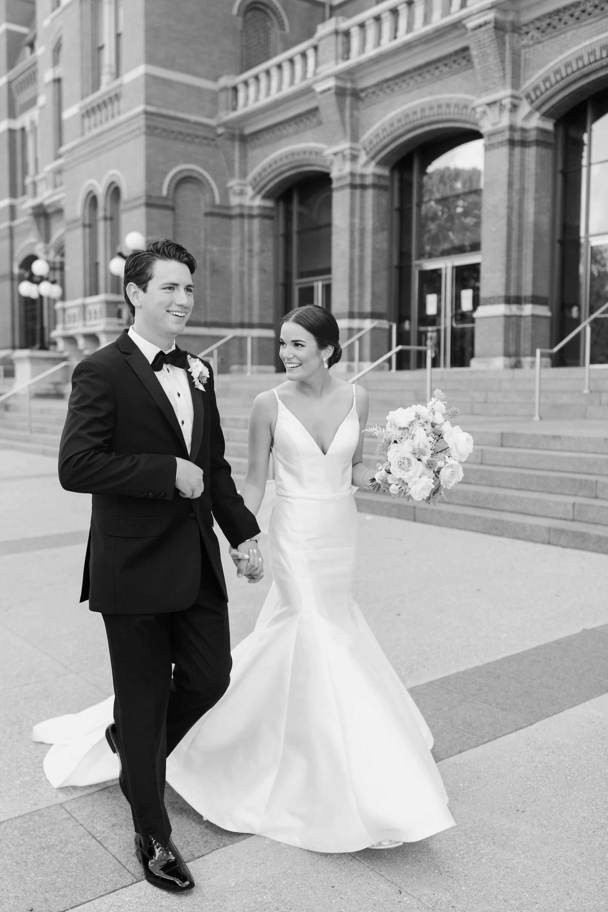 A bride and groom walking hand in hand outside a historic brick building, dressed in wedding attire. The bride is holding a bouquet and wearing a white wedding gown, while the groom is in a black tuxedo with a bow tie. Both are smiling.