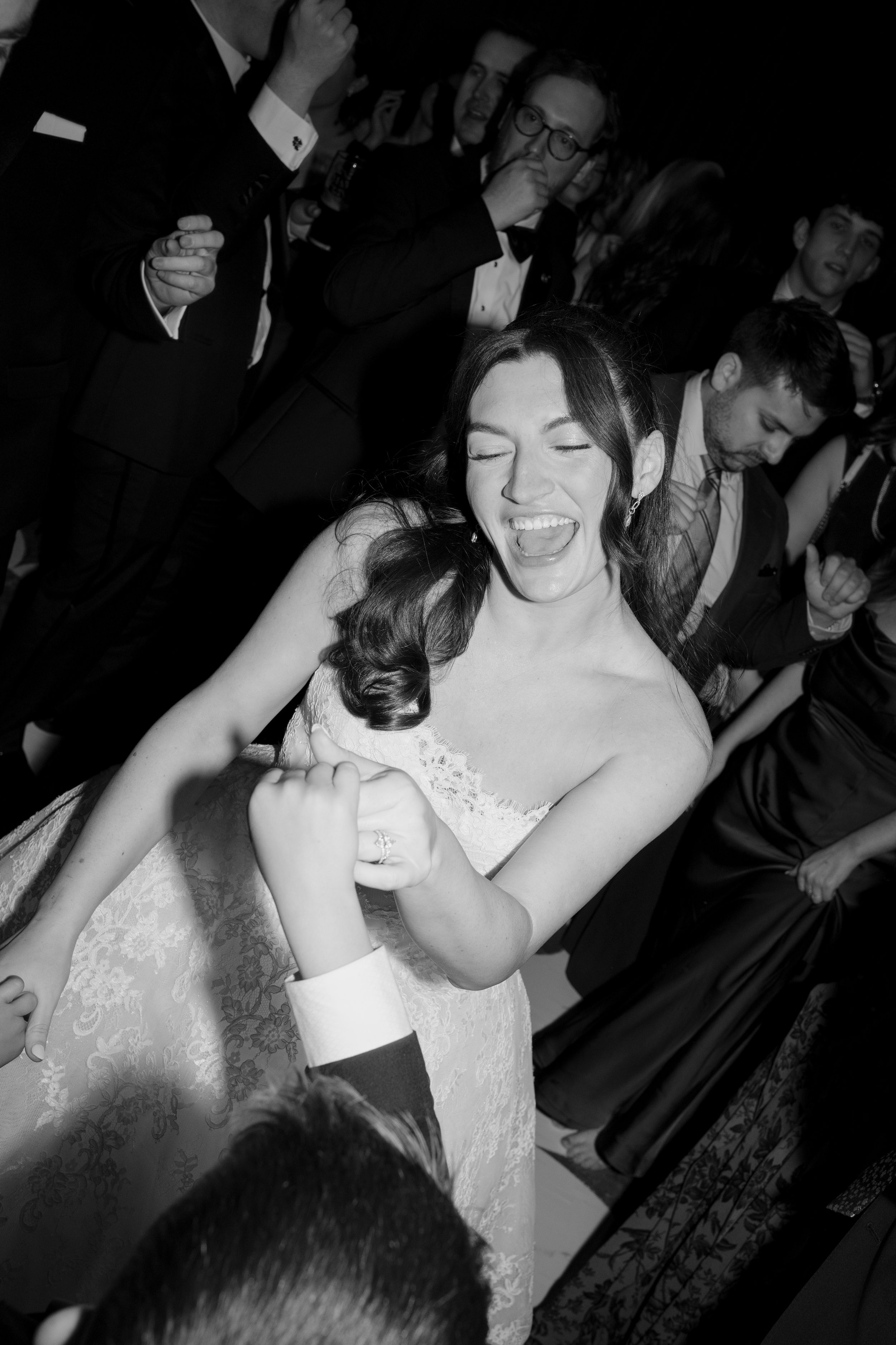 A group of people at a formal event, with a woman in a wedding dress in the foreground smiling and laughing while holding hands with a man, surrounded by others in tuxedos and dresses.