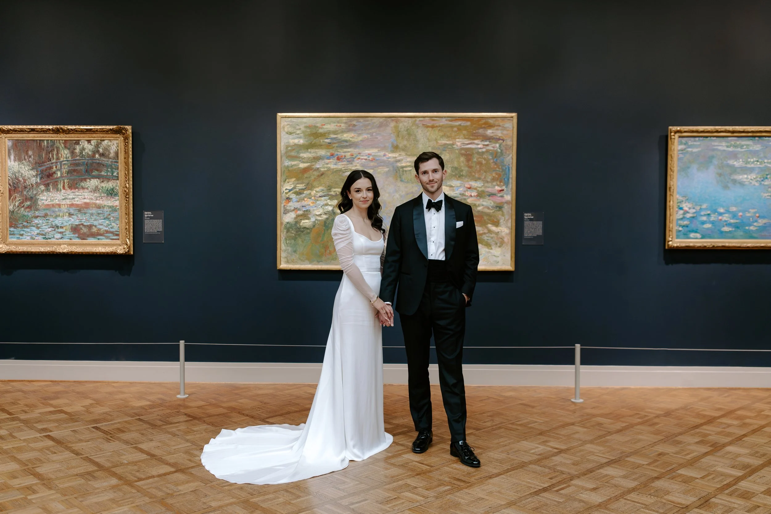 A couple dressed in formal wedding attire standing in an art gallery with paintings on dark wall behind them.