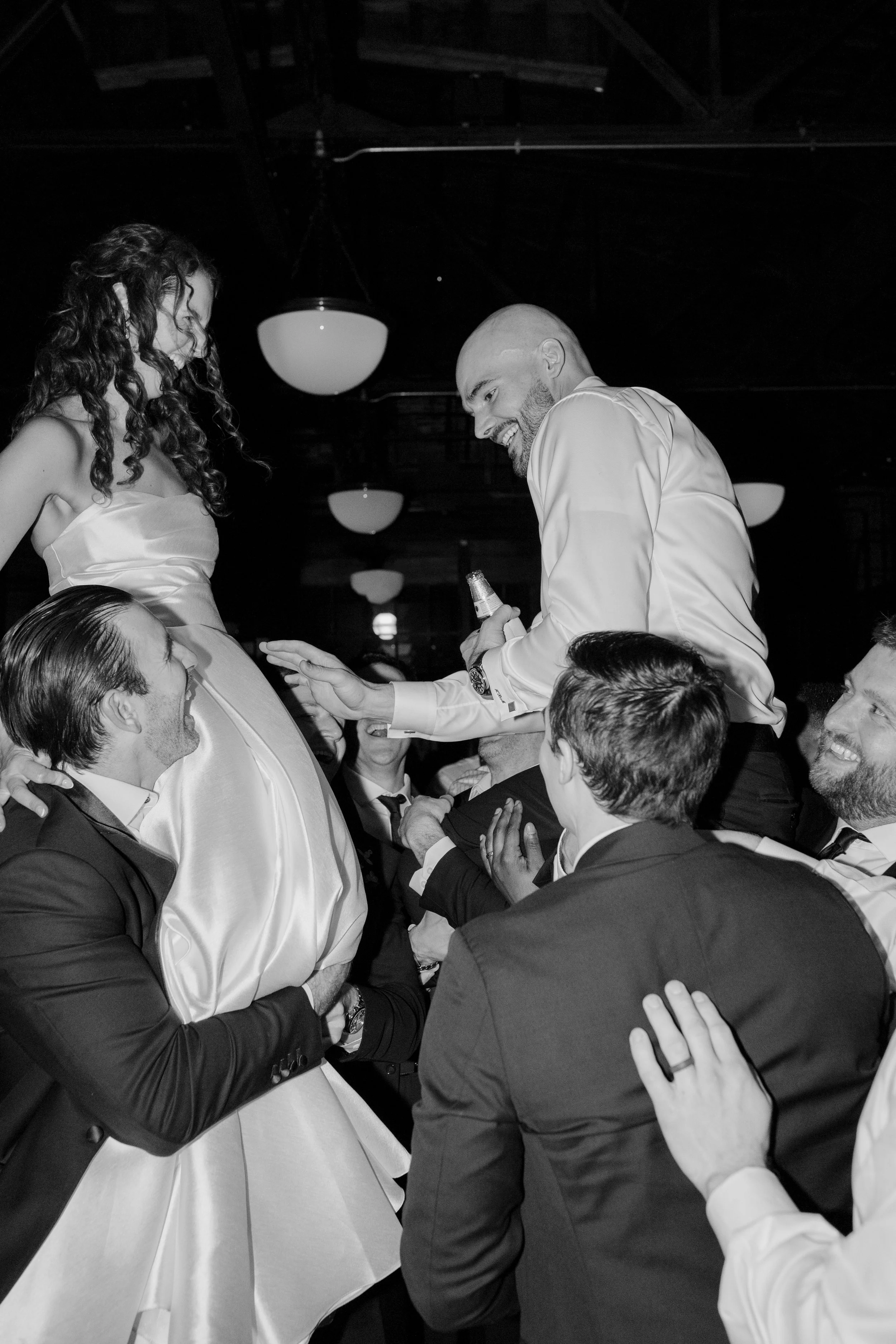A group of people in formal attire at a celebration, with a woman in a satin dress and a man in a shirt and tie smiling, engaging with others around. The setting appears to be indoors with ceiling lights visible.