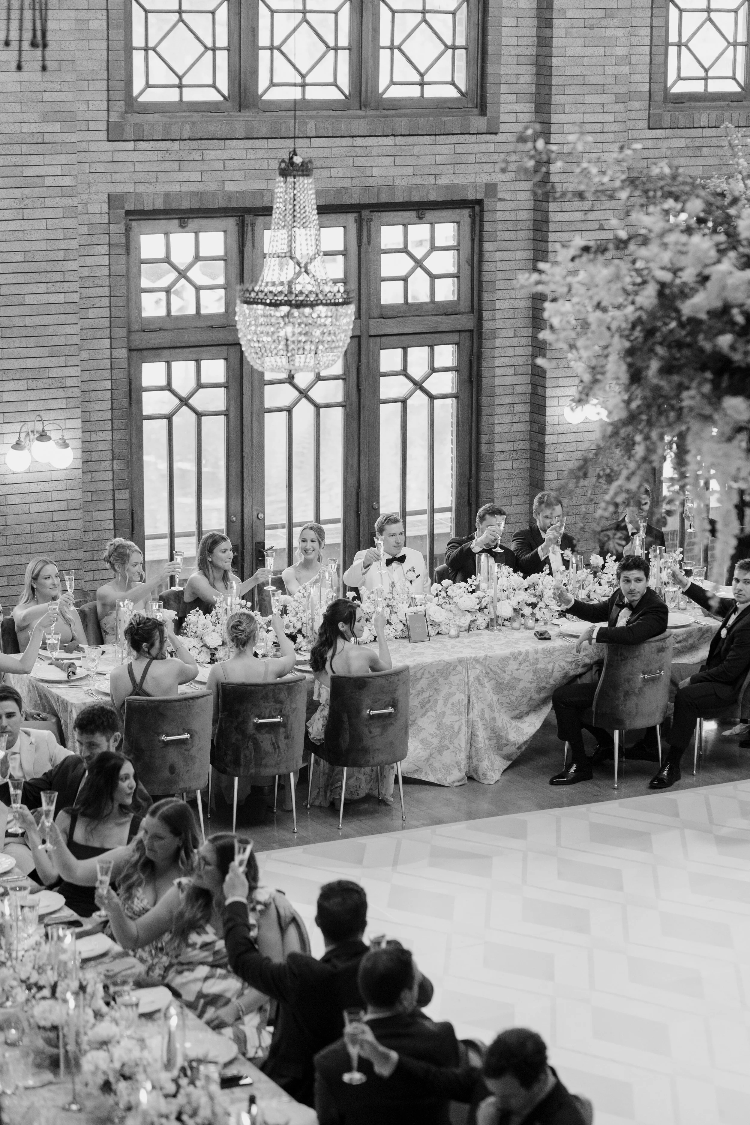 Guests at a formal wedding reception raise champagne glasses for a toast inside a brick-walled venue with large decorative windows and a chandelier.