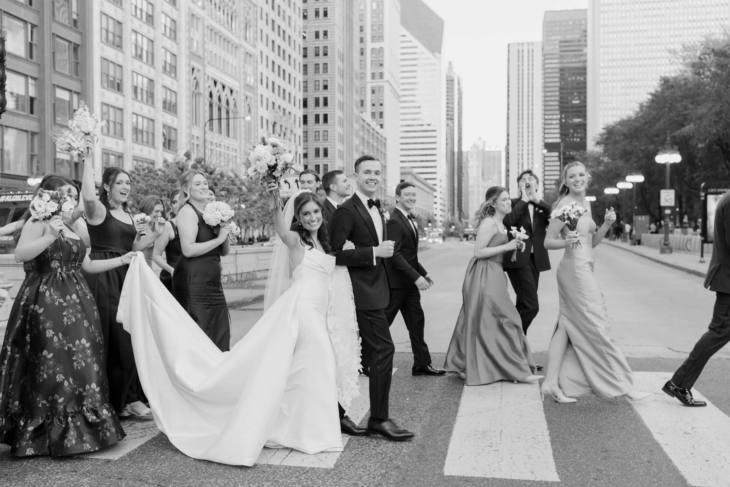 Black and white photo of a wedding party crossing a city street, city skyscrapers in the background, the bride and groom smiling, surrounded by bridesmaids and groomsmen holding flowers and dressed in formal attire.