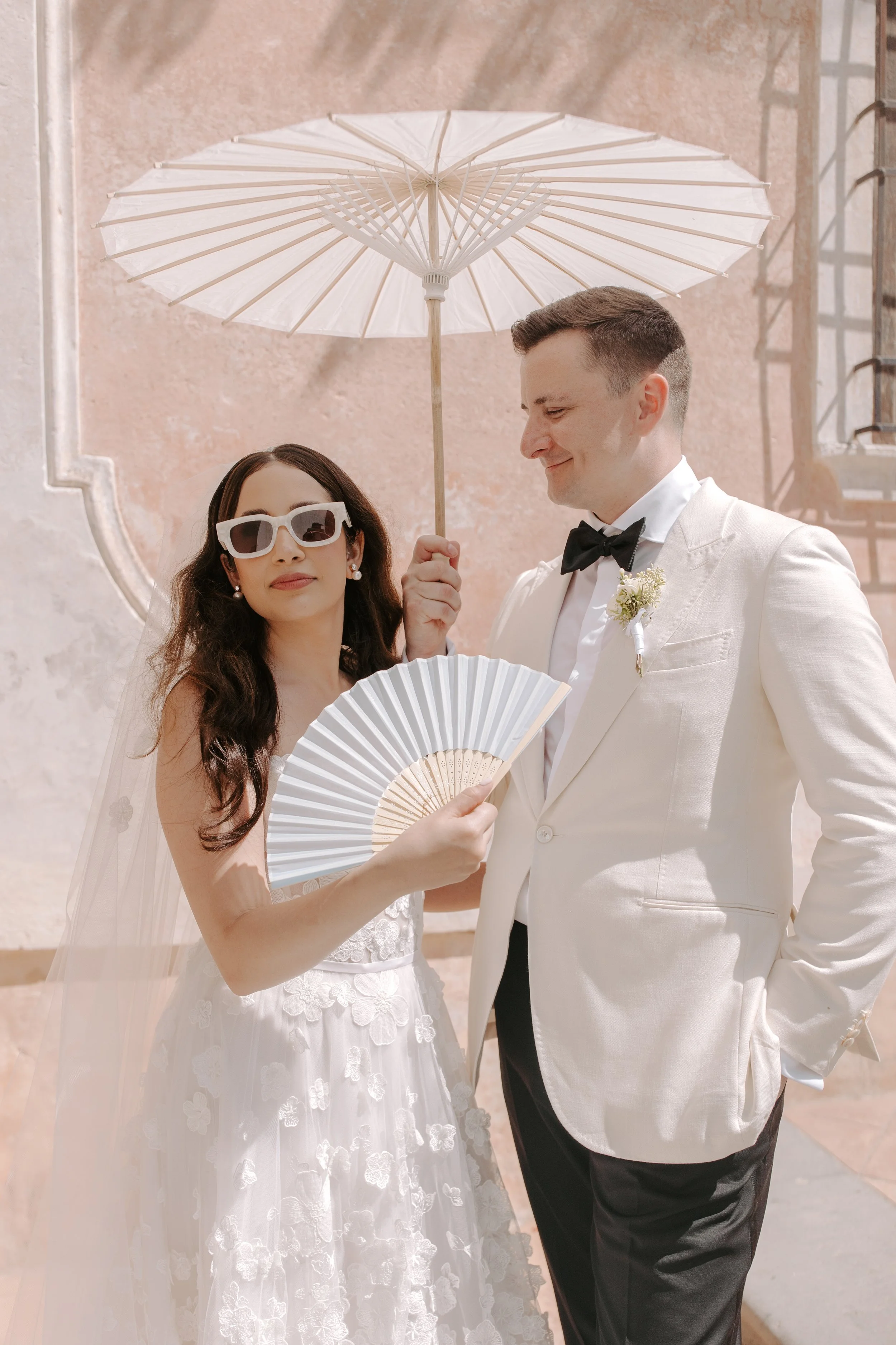 A bride and groom dressed in wedding attire standing outdoors under a white parasol, with the bride holding a white fan and wearing white sunglasses, on a sunny day.