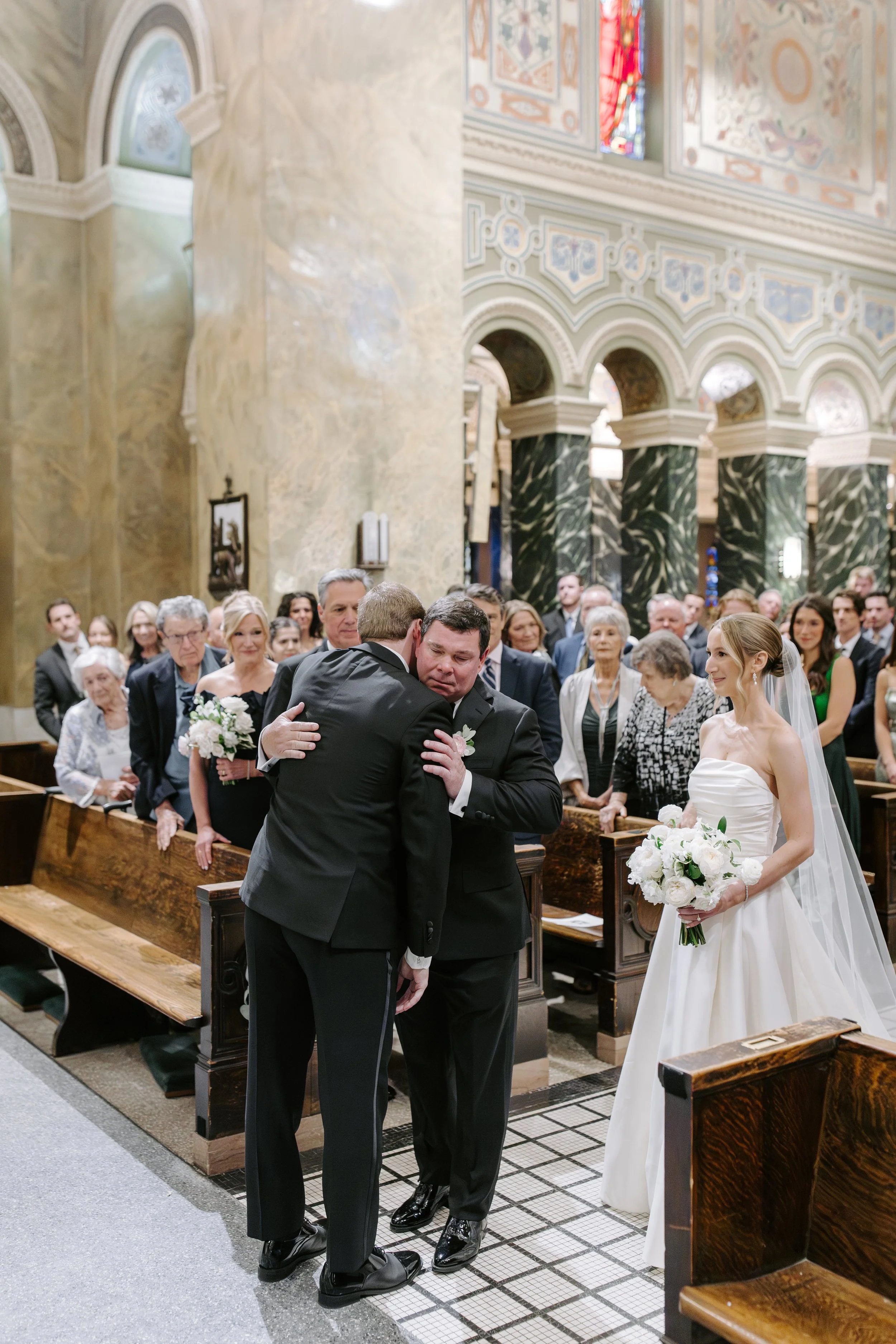 A wedding ceremony inside a church with a bride holding a bouquet of white flowers, standing near the altar, and two men hugging at the front. Guests are seated in pews and the church features ornate archways, stained glass windows, and decorative wa