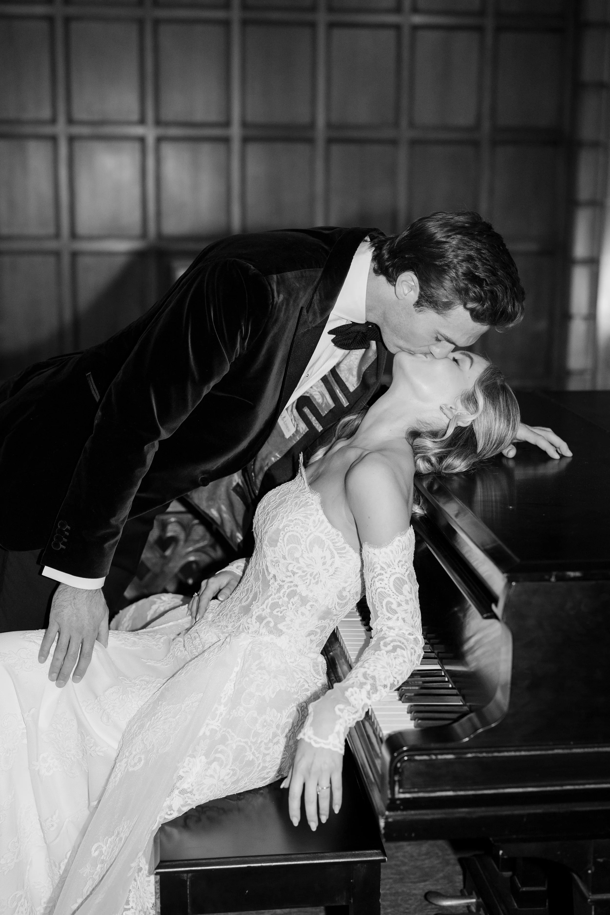 A man in a tuxedo leaning over a woman in a lace wedding dress as they share a kiss by a piano.