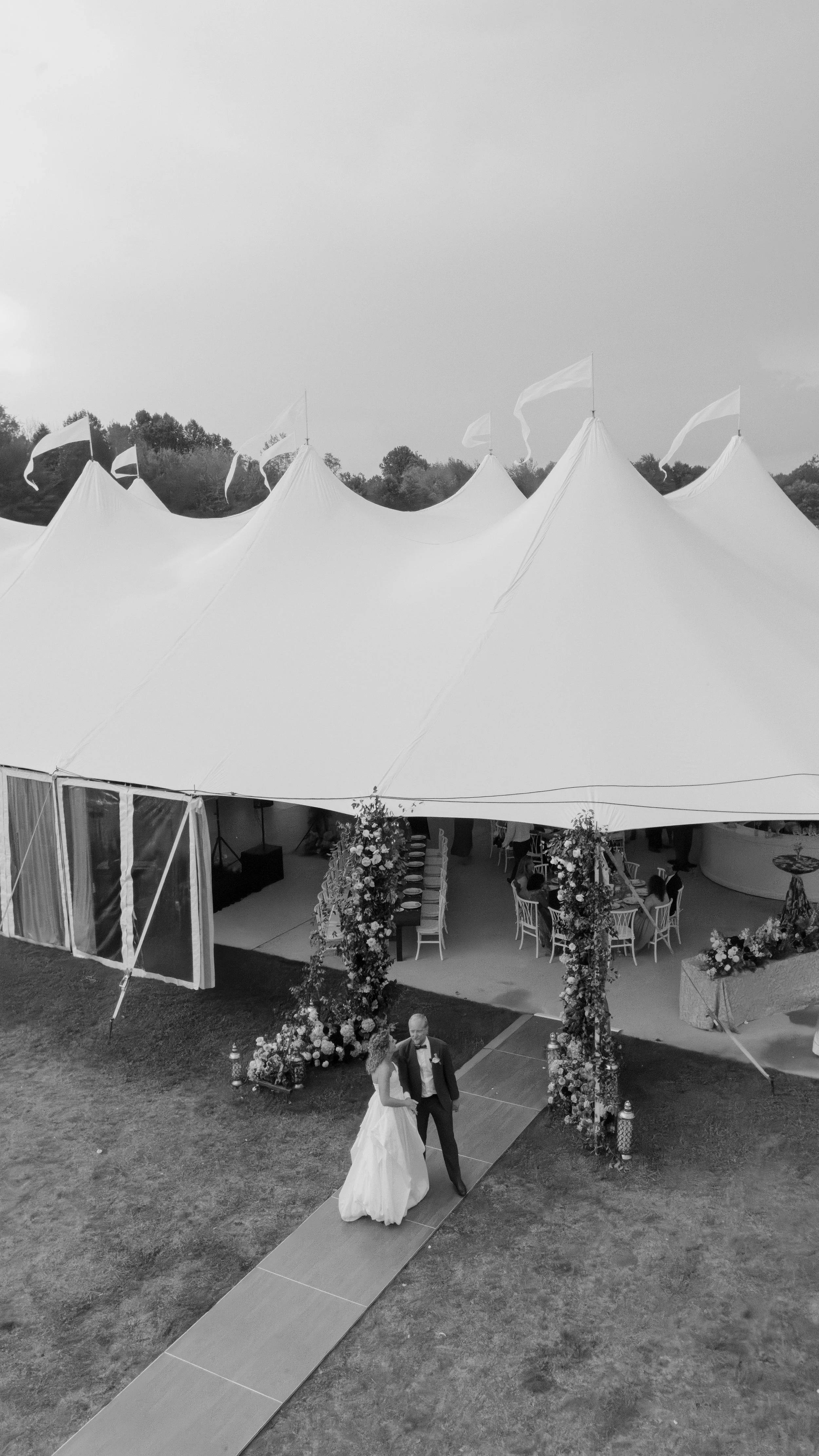 A bride and groom walk together on a wedding aisle under a large tent decorated with flowers. The tent is set up in a grassy outdoor area, and guests are seated at tables inside.