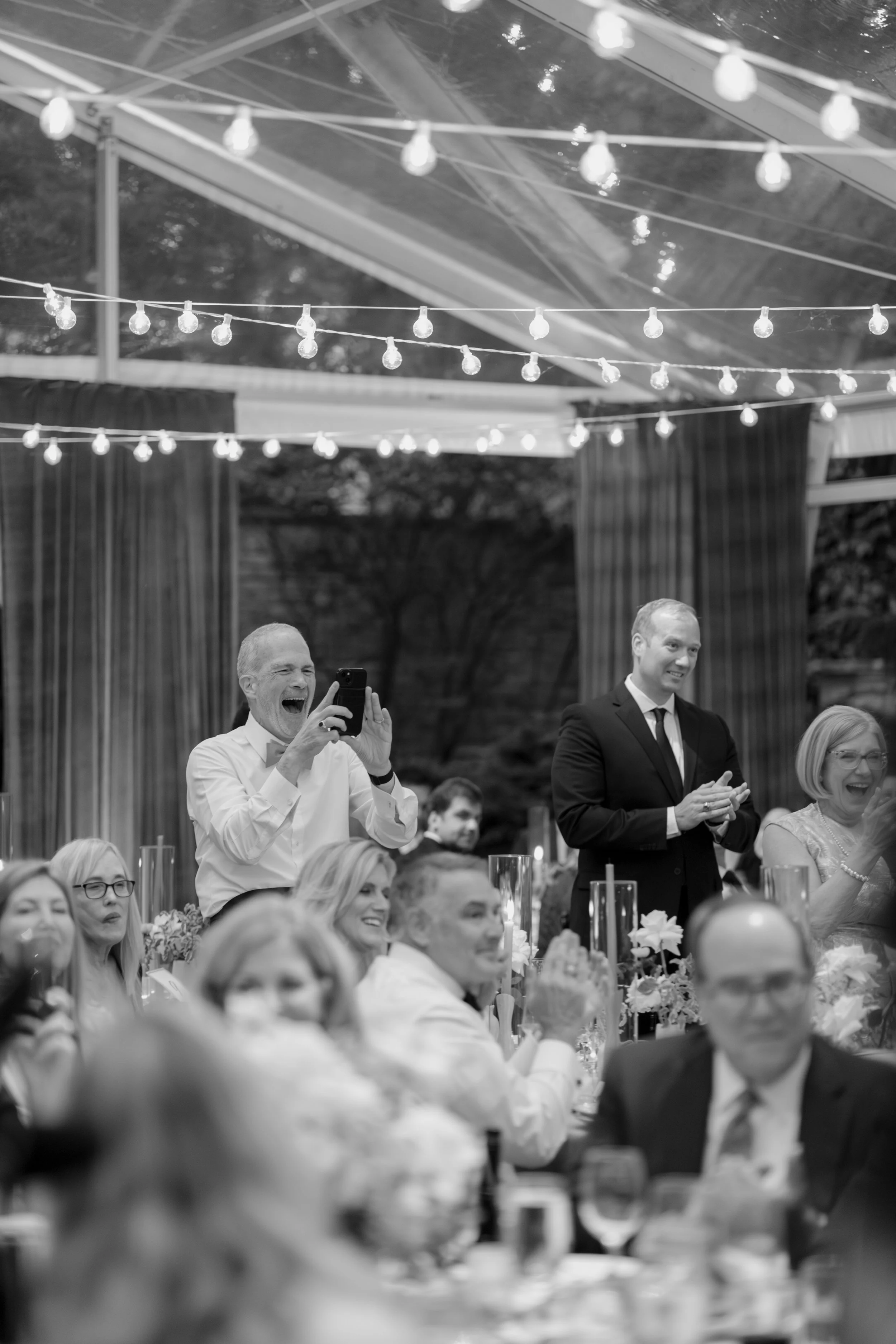 People at a formal event or celebration, including a man in a tuxedo and a woman with glasses, seated at a decorated table with candles and flowers. String lights are hanging above inside a tent or reception hall.