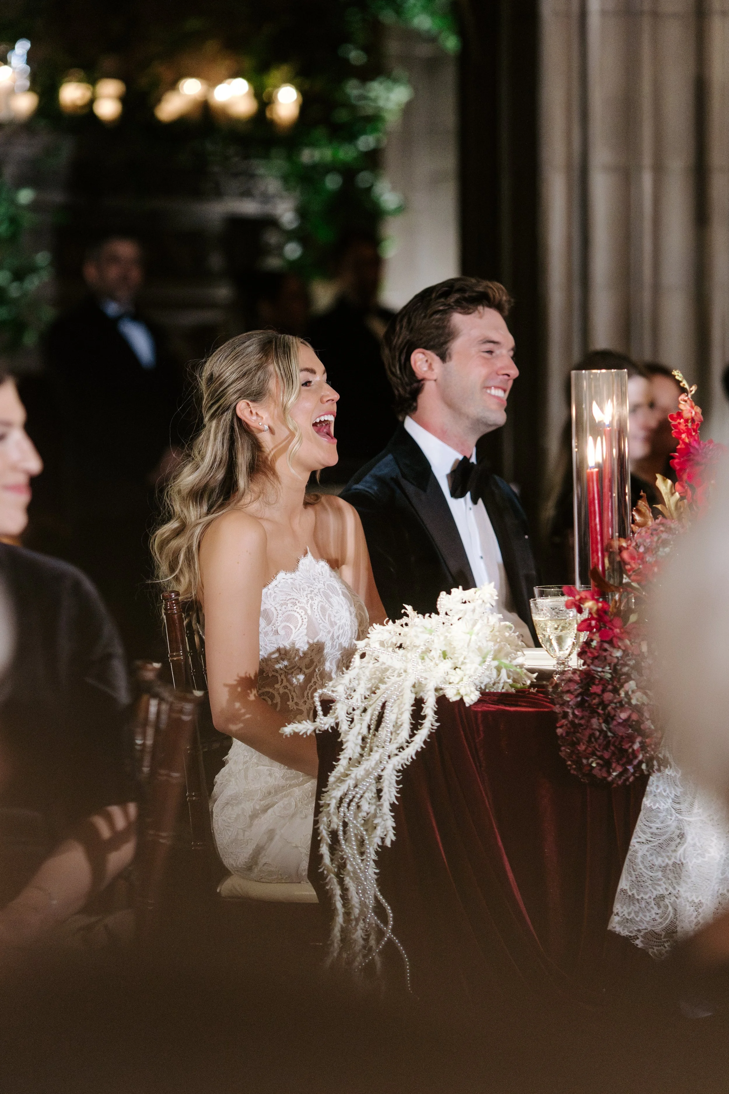 A bride and groom smiling at their wedding reception, sitting at a table decorated with flowers and candles.
