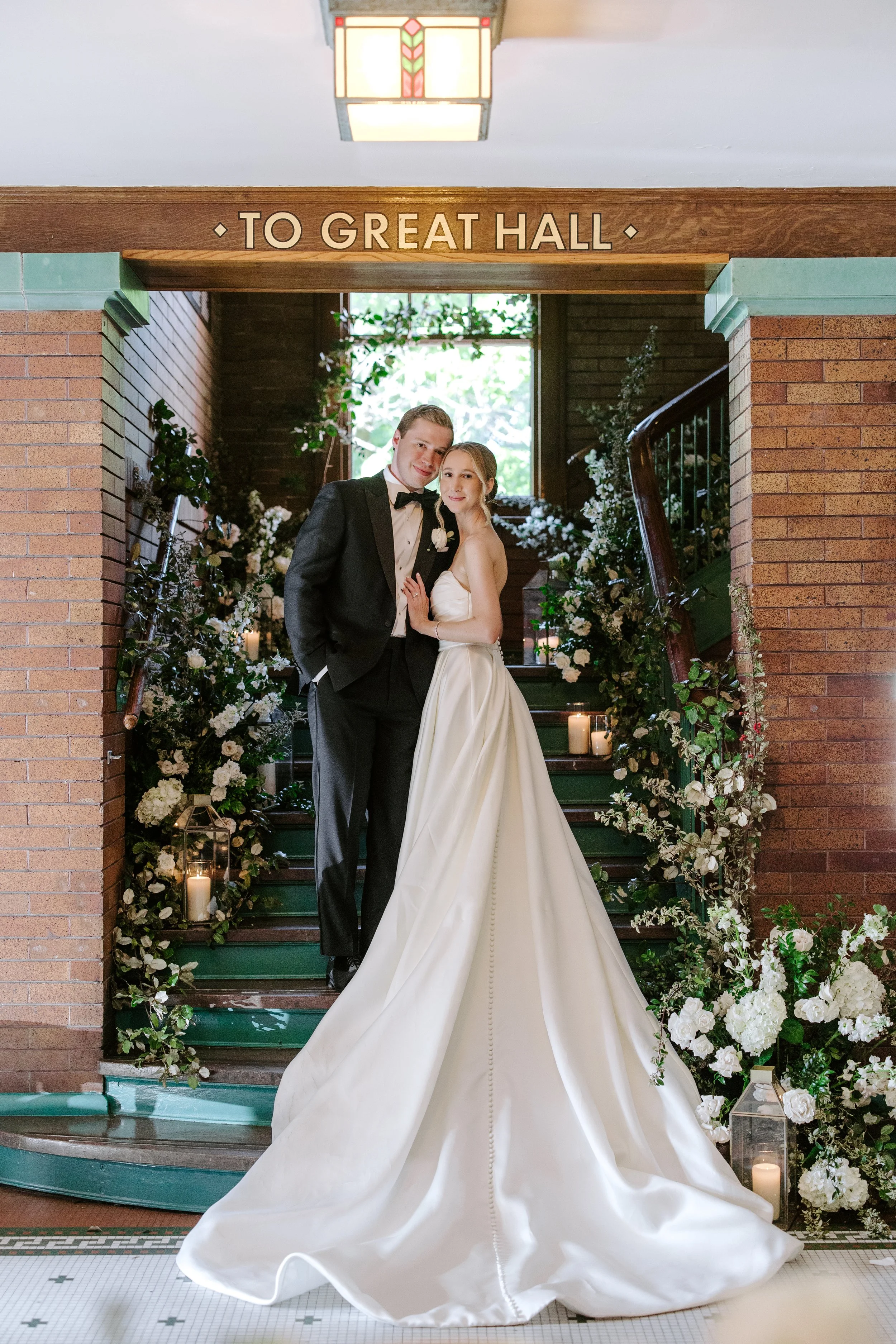 A bride and groom standing on a staircase decorated with white flowers and candles, inside a building with exposed brick walls and a stained glass ceiling, under a wooden sign that reads 'TO GREAT HALL'.