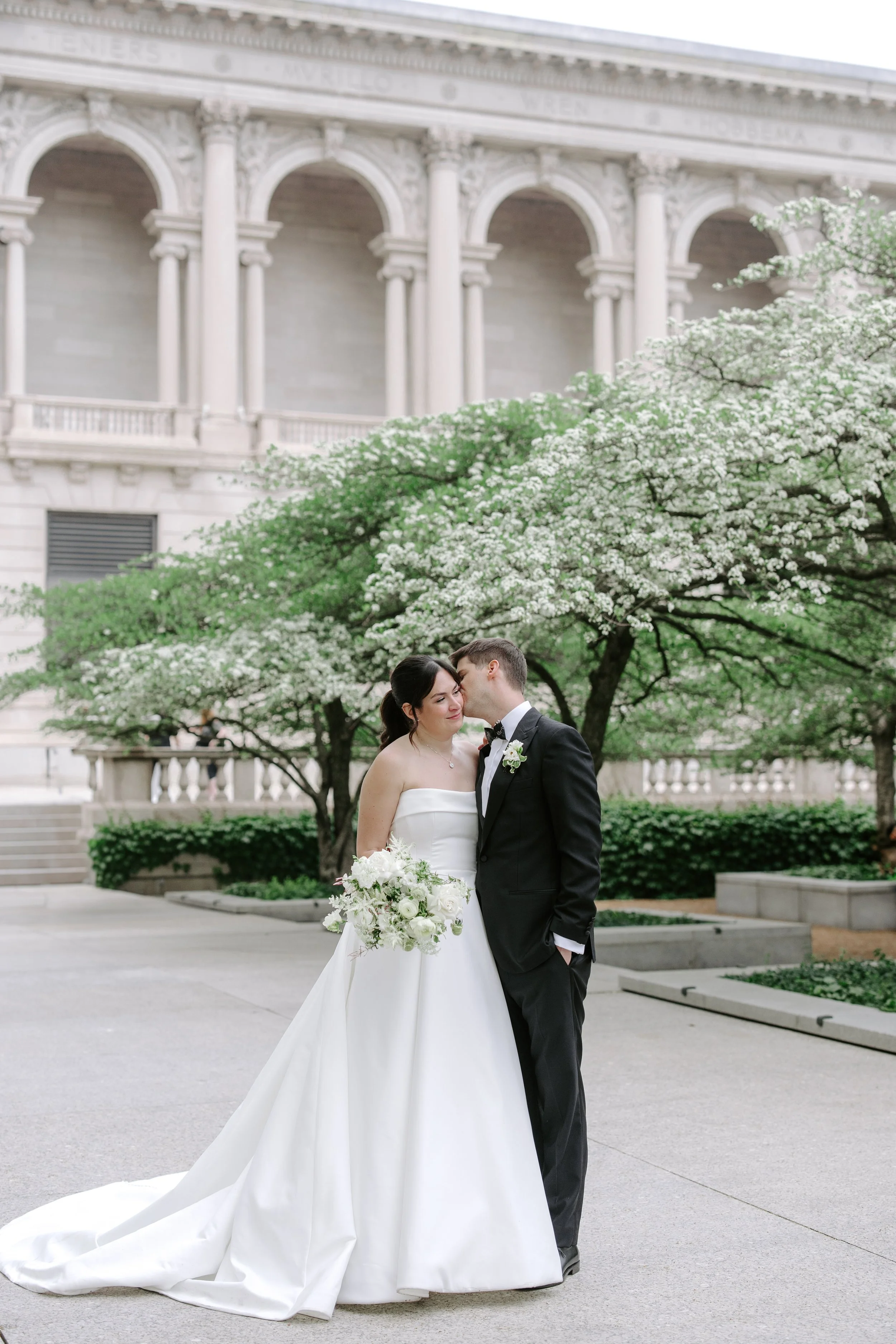 A bride and groom embrace outdoors in front of a flowering tree and a historic building with columns and arches, during a wedding ceremony.