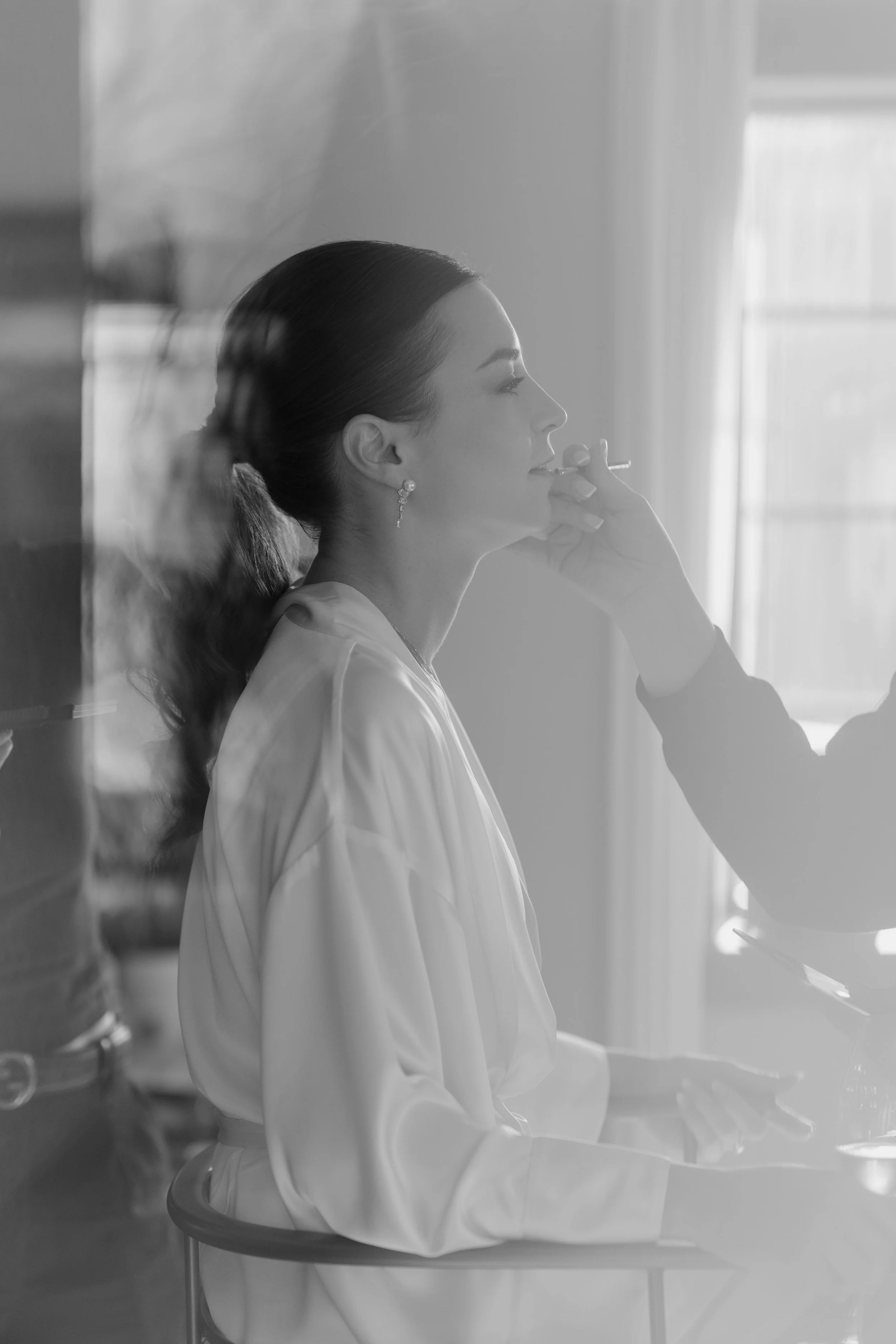 A black and white photo of a woman with dark hair in a ponytail, sitting at a table, smoking a cigarette, with a window in the background.