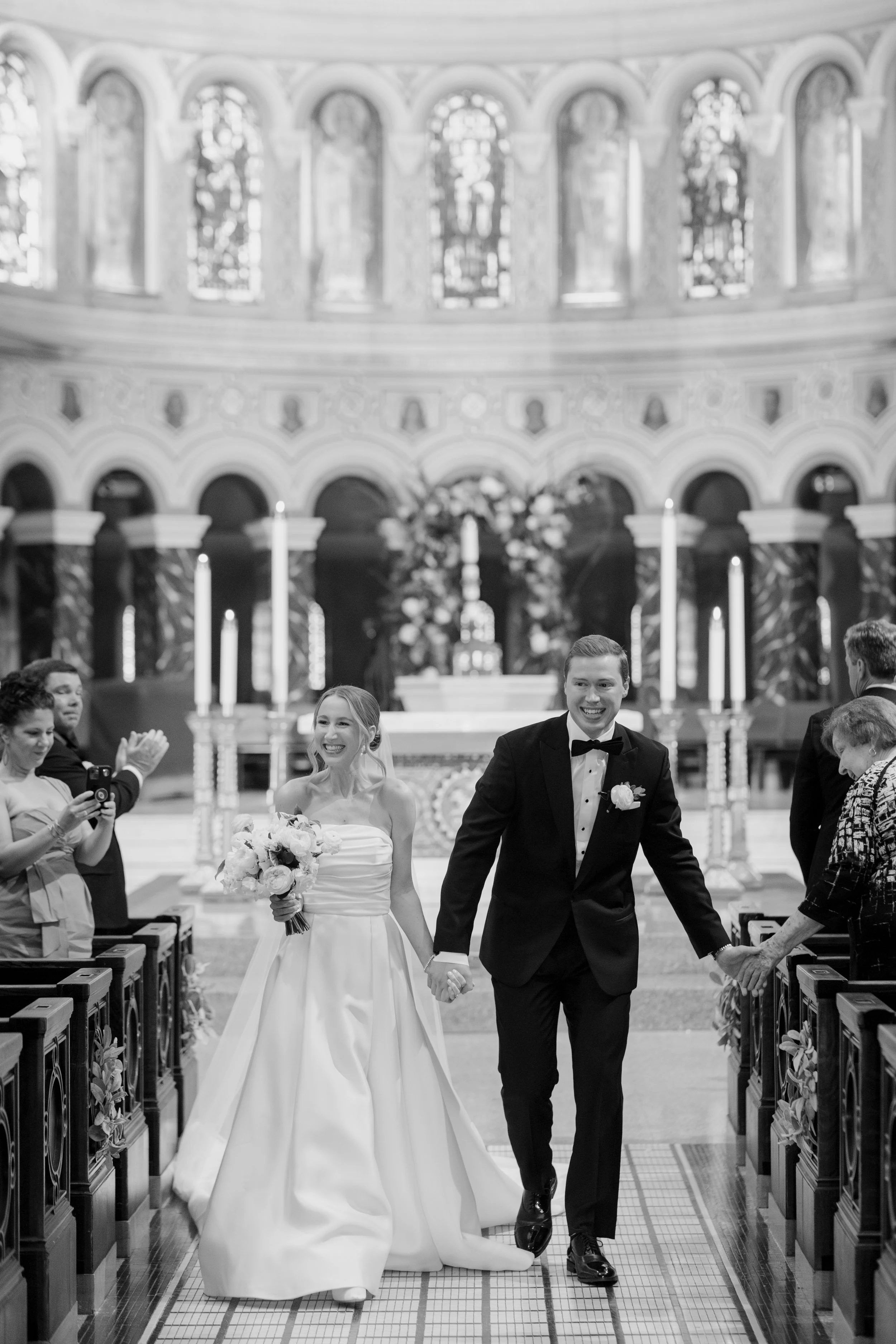 A bride and groom holding hands and walking down the aisle in a church with guests on either side. The bride is in a white wedding gown holding a bouquet, and the groom is in a black tuxedo with a bow tie. The church has high ceilings, large stained 