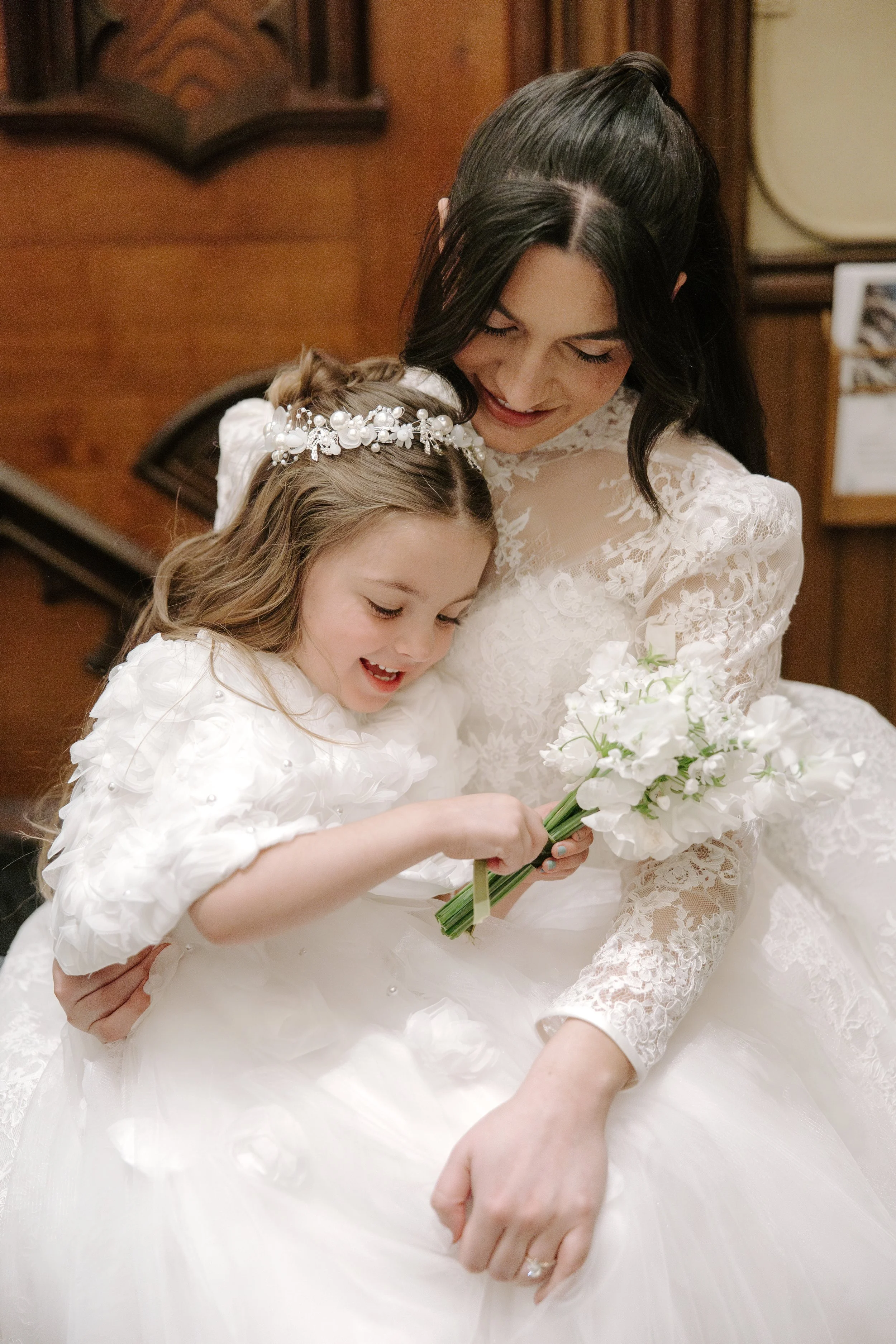 A woman in a lace wedding dress hugs a young girl in a white dress with a pearl and floral headband, holding a bouquet of white flowers, in a room with wood-paneled walls.