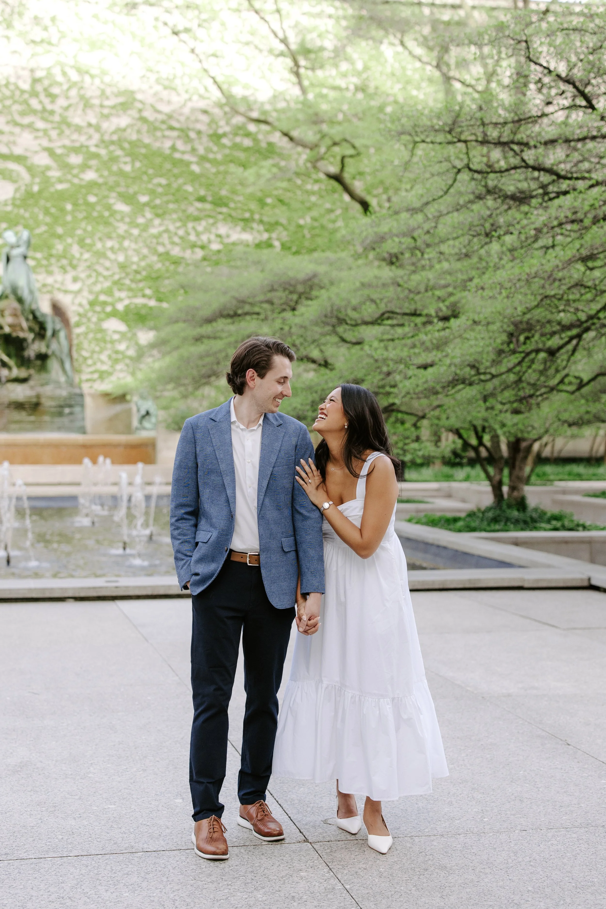 A happy couple in formal attire standing hand in hand outdoors near a fountain, surrounded by green trees, smiling and looking at each other.